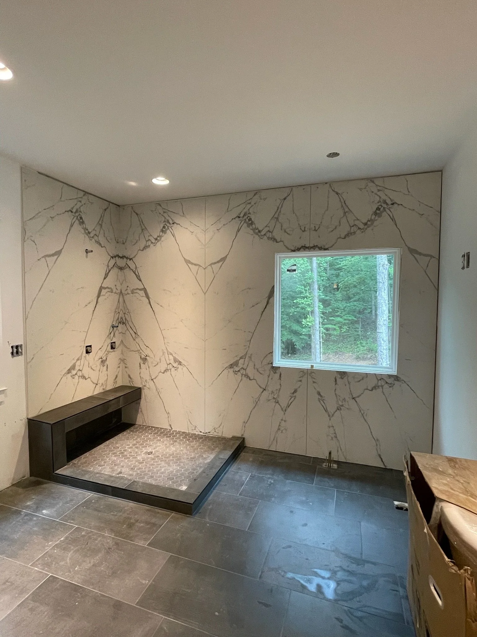 An unfinished room under construction with marble-patterned wall tiles, a window showing a wooded outdoor view, and a low black platform for a shower or fireplace. The floor has large dark tiles, and there is a cardboard box on the right side.