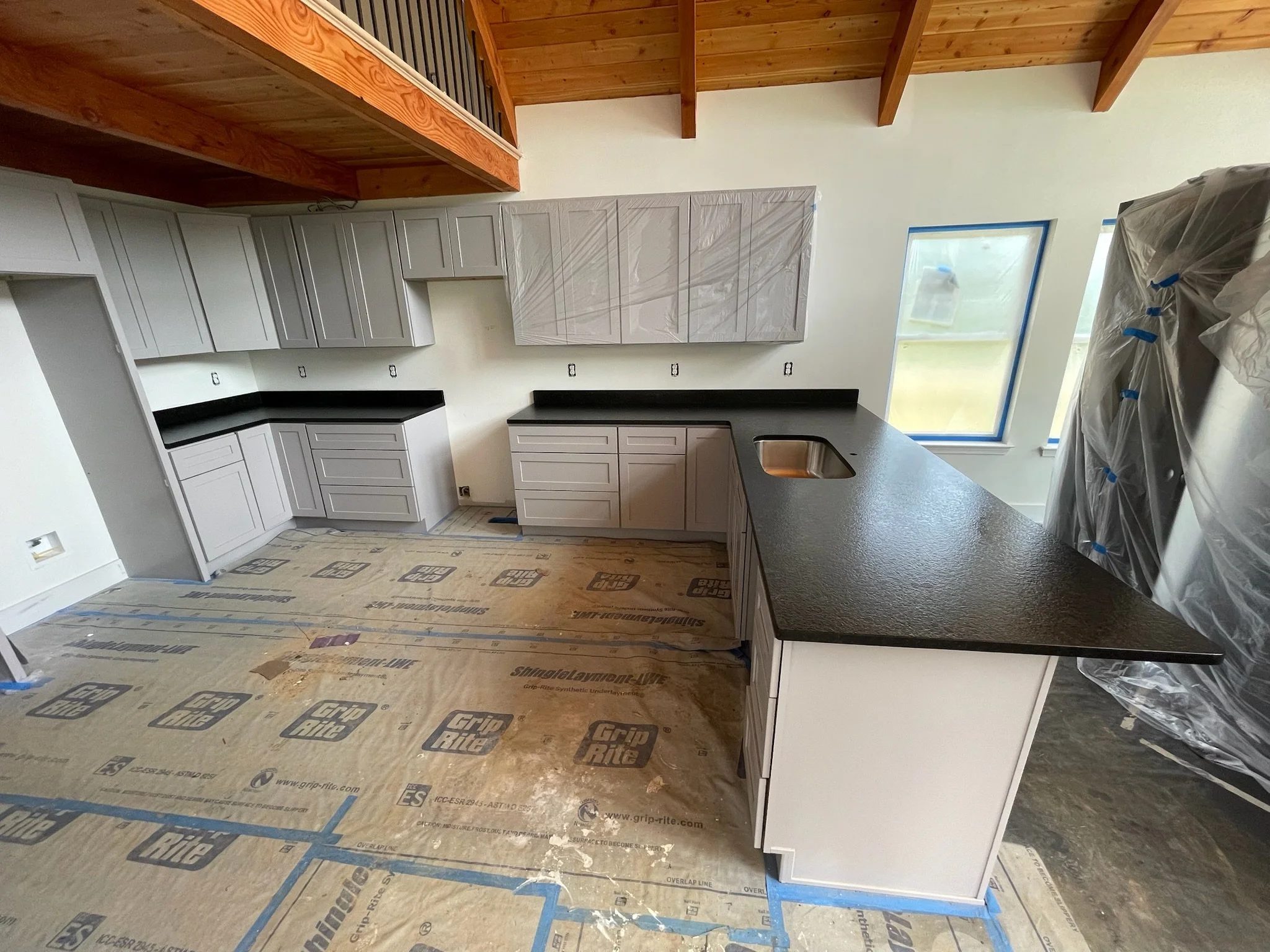 Kitchen under construction with gray cabinets, black countertops, and an island with a sink. Plastic protective covering on appliances and a wooden ceiling.