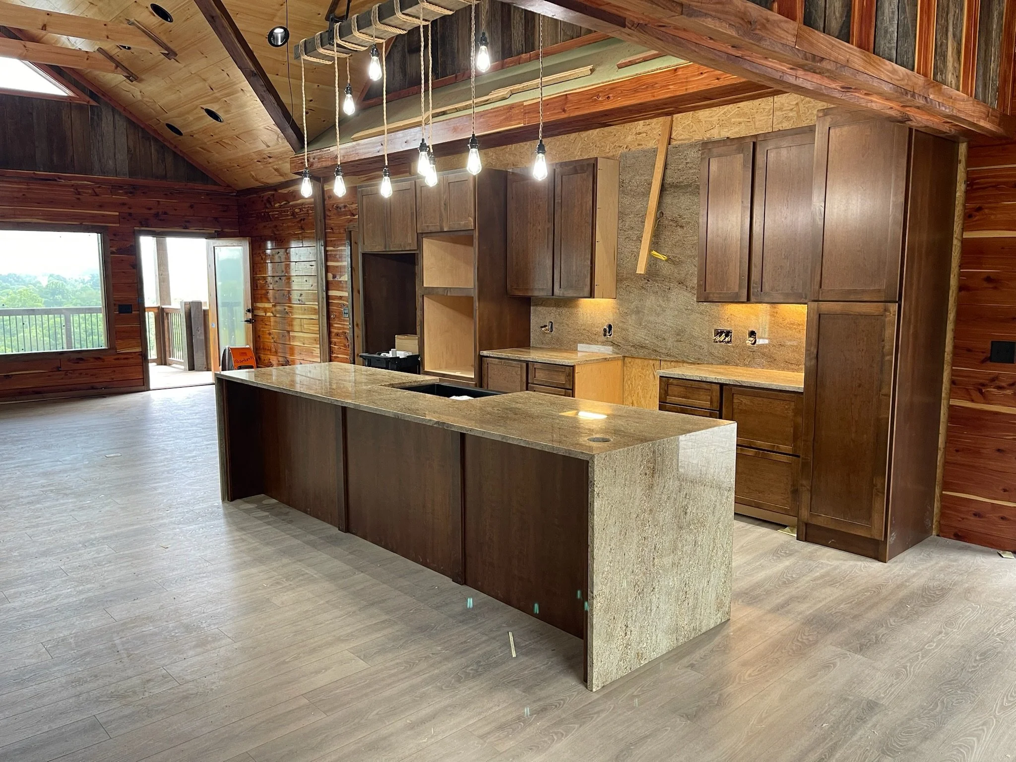 Modern kitchen with an island, wooden cabinets, and hardwood floors featuring exposed beams and large windows in a rustic home.