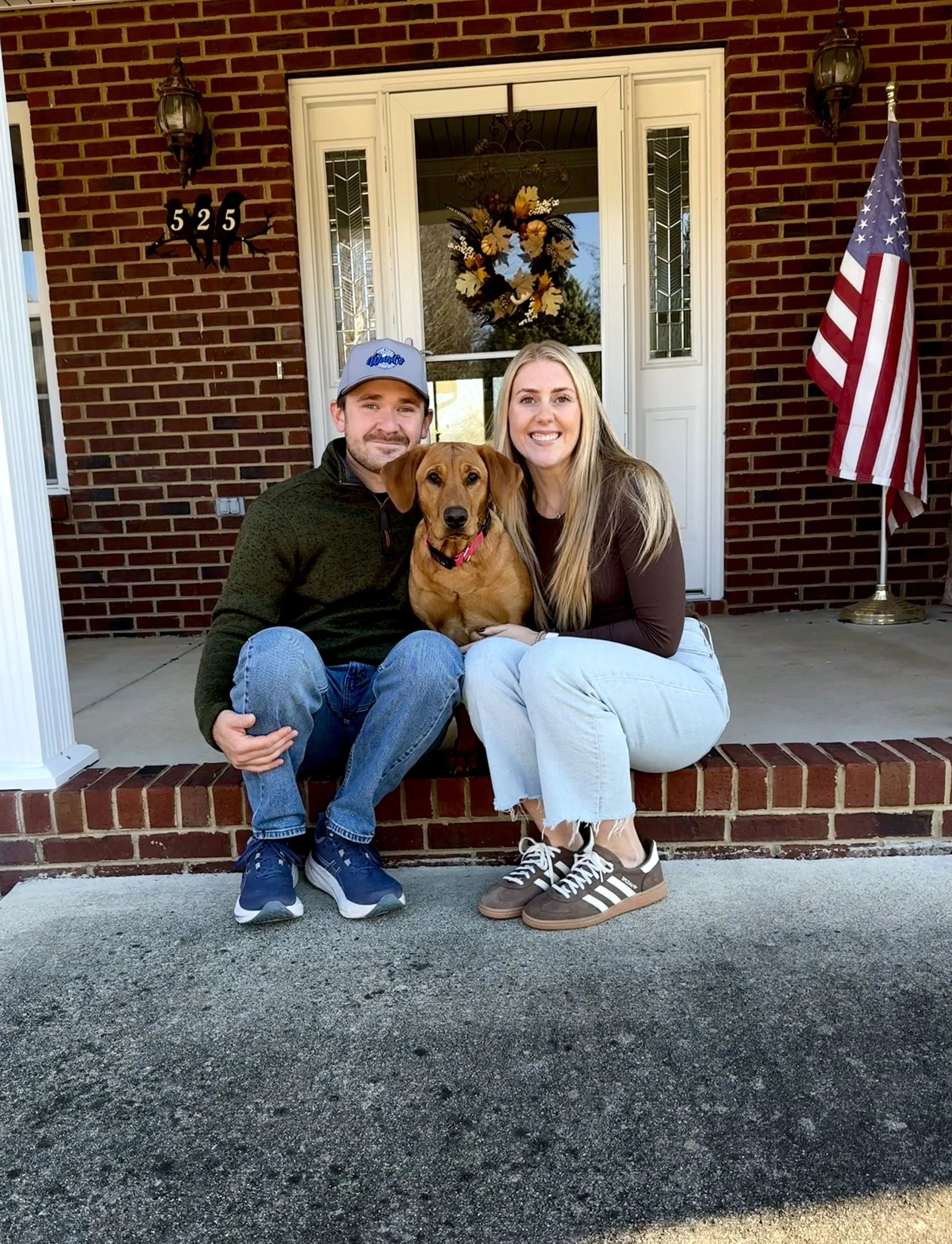 A smiling man, woman, and a dog sitting on the front porch of a brick house decorated with an American flag, a floral wreath on the door, and house numbers 525.