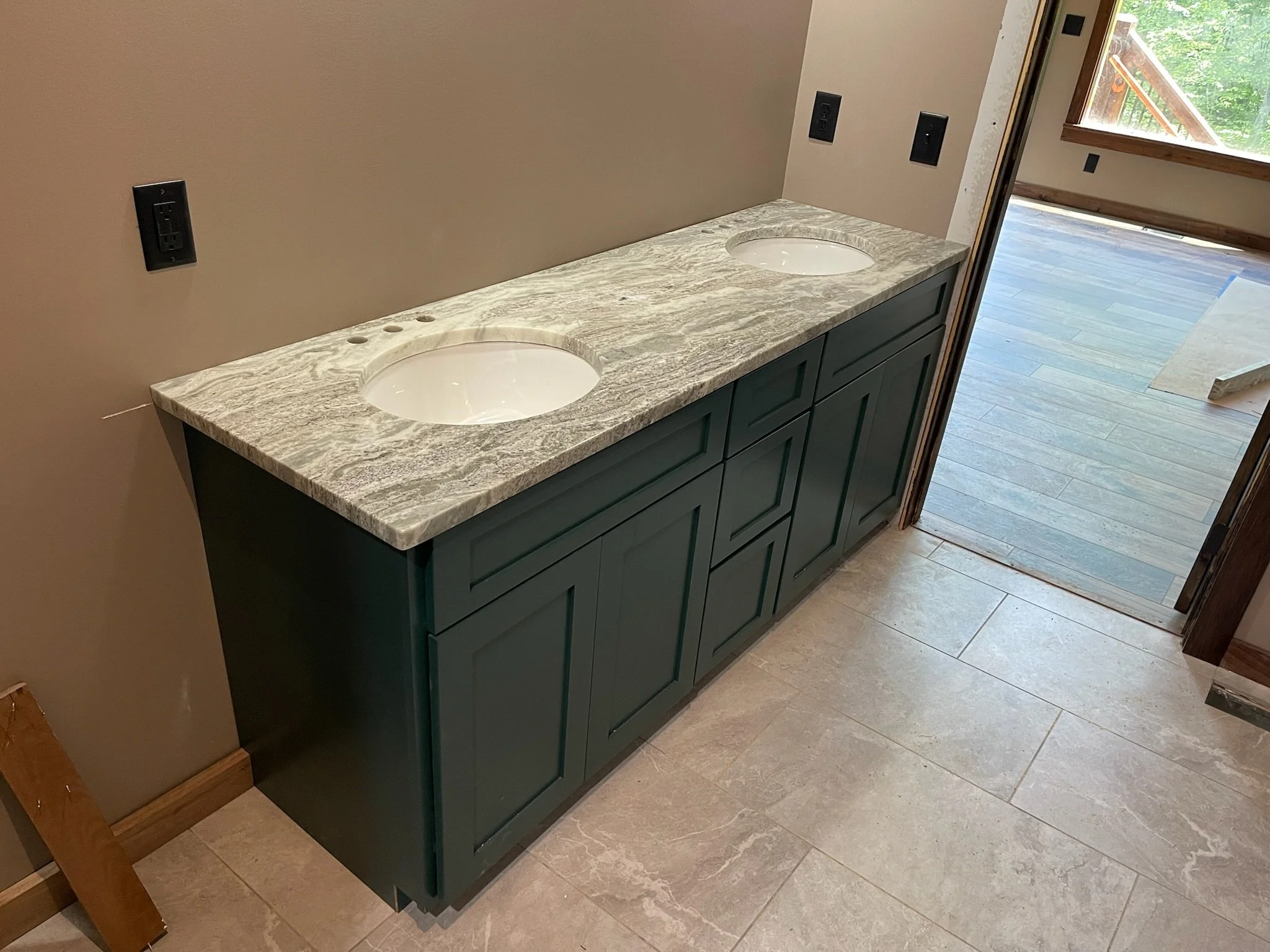 A bathroom vanity with a gray, marbled countertop and dual sinks, set against a beige wall with electrical outlets, with a view of a wooden deck outside through an open door.