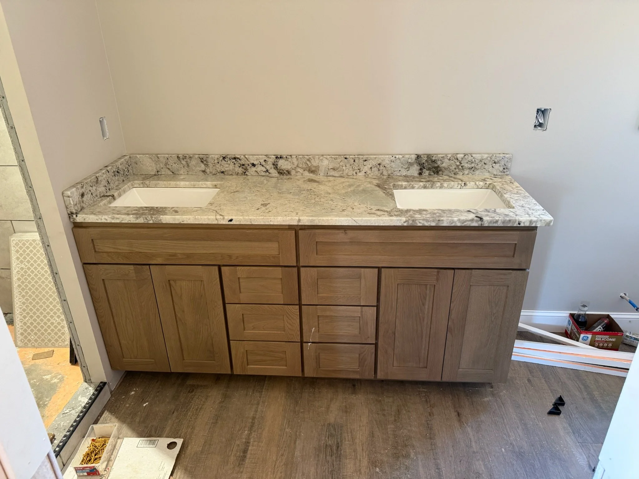Interior view of a bathroom or laundry room with a wooden cabinet with a granite countertop, two undermount sinks, and electrical outlets without covers.