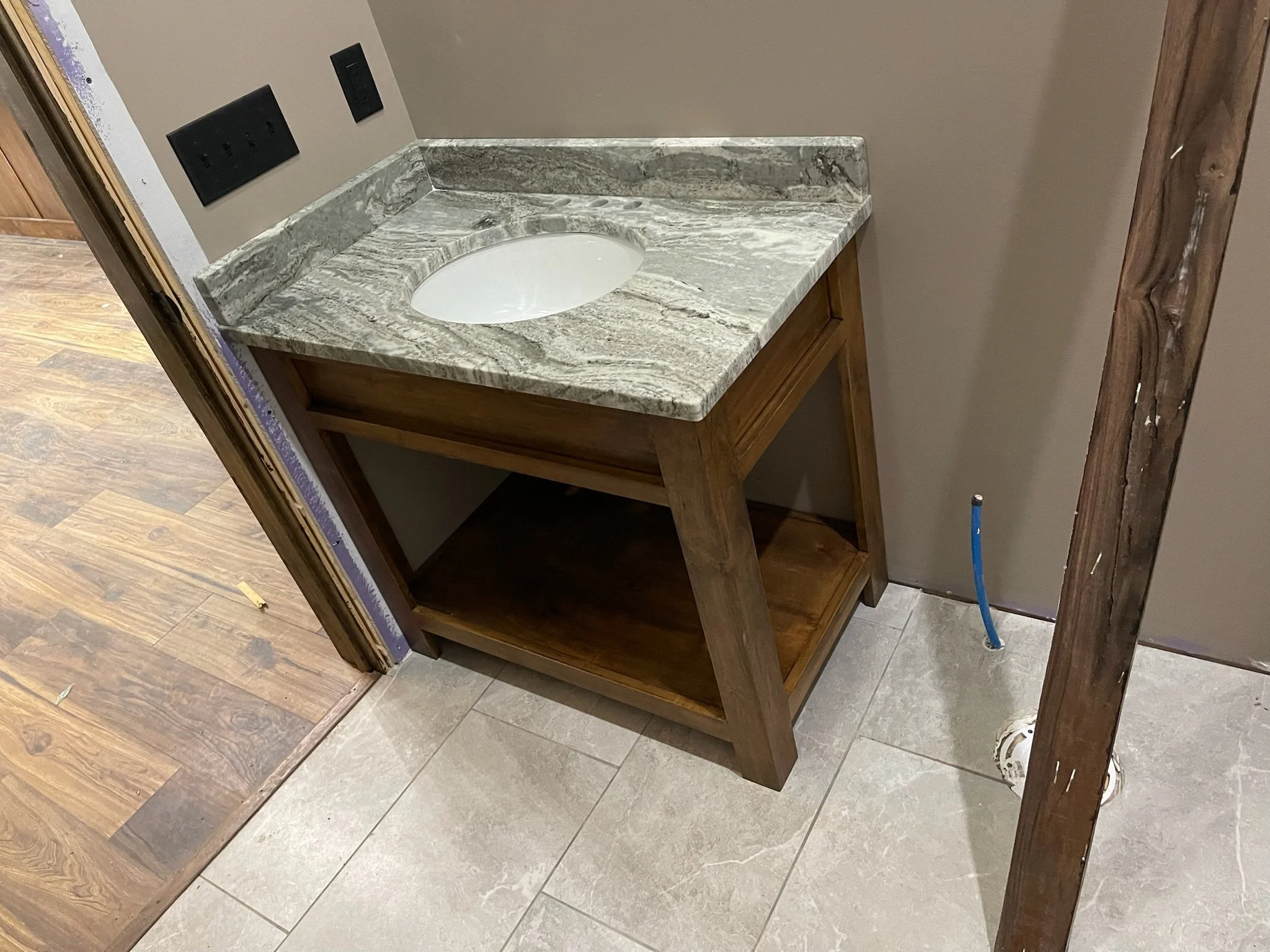 A bathroom vanity with a marble countertop and an oval sink, without a faucet, and an open bottom shelf, next to unfinished wall and plumbing.