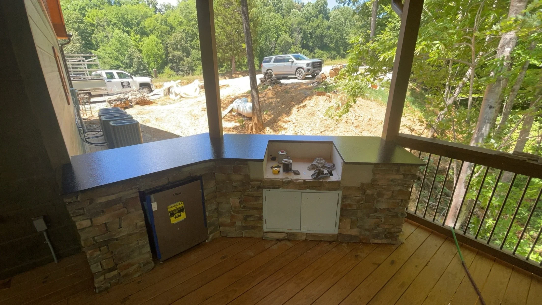 View from a porch with a black countertop, brick base, and a small storage cabinet, overlooking a construction site with two parked trucks and trees in the background.