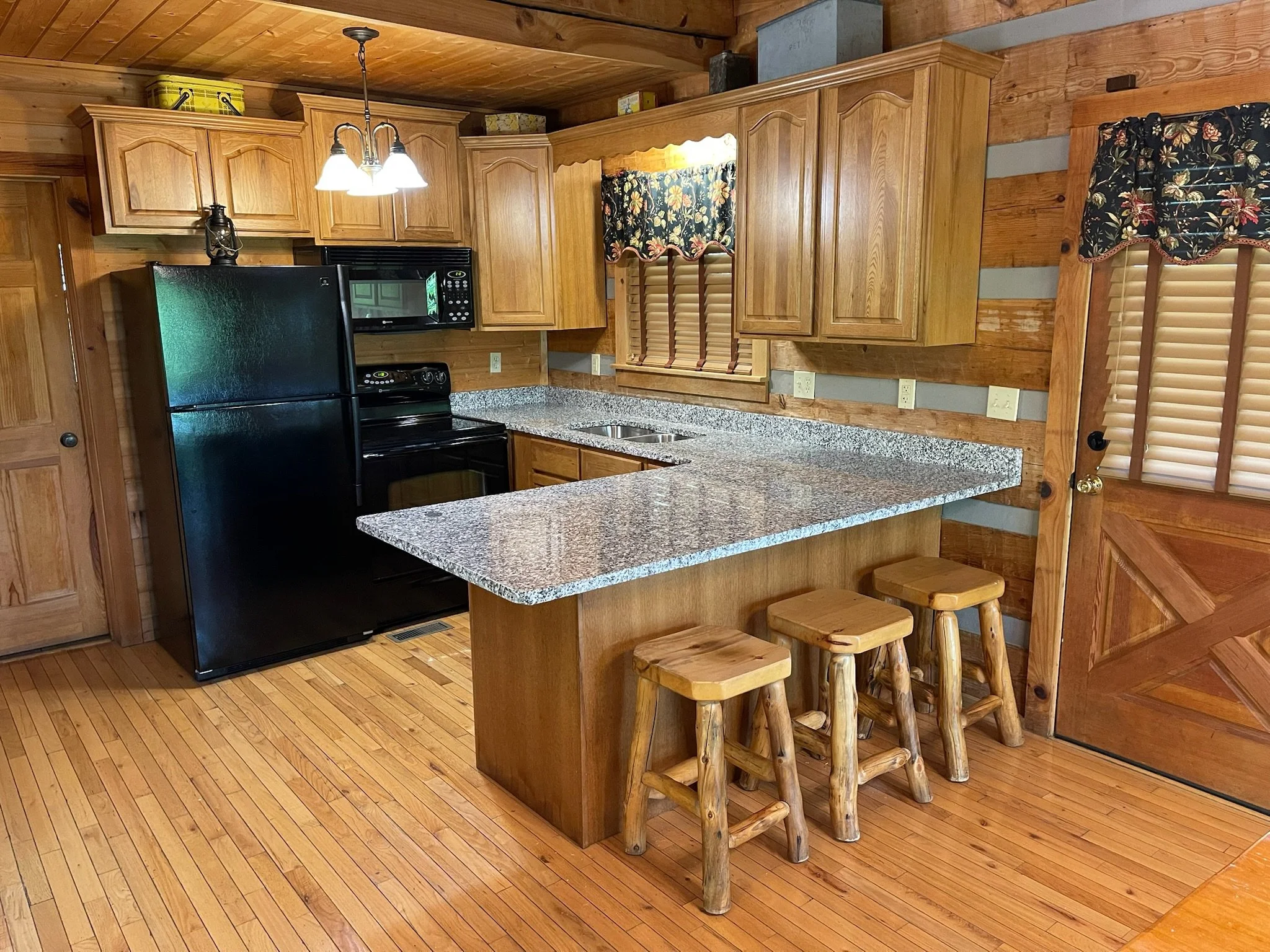 A rustic kitchen with wooden cabinets, granite countertop, black appliances including a refrigerator, microwave, and oven. There is a small north turned window with floral curtains, a wooden door with blinds, and a kitchen island with three wooden st
