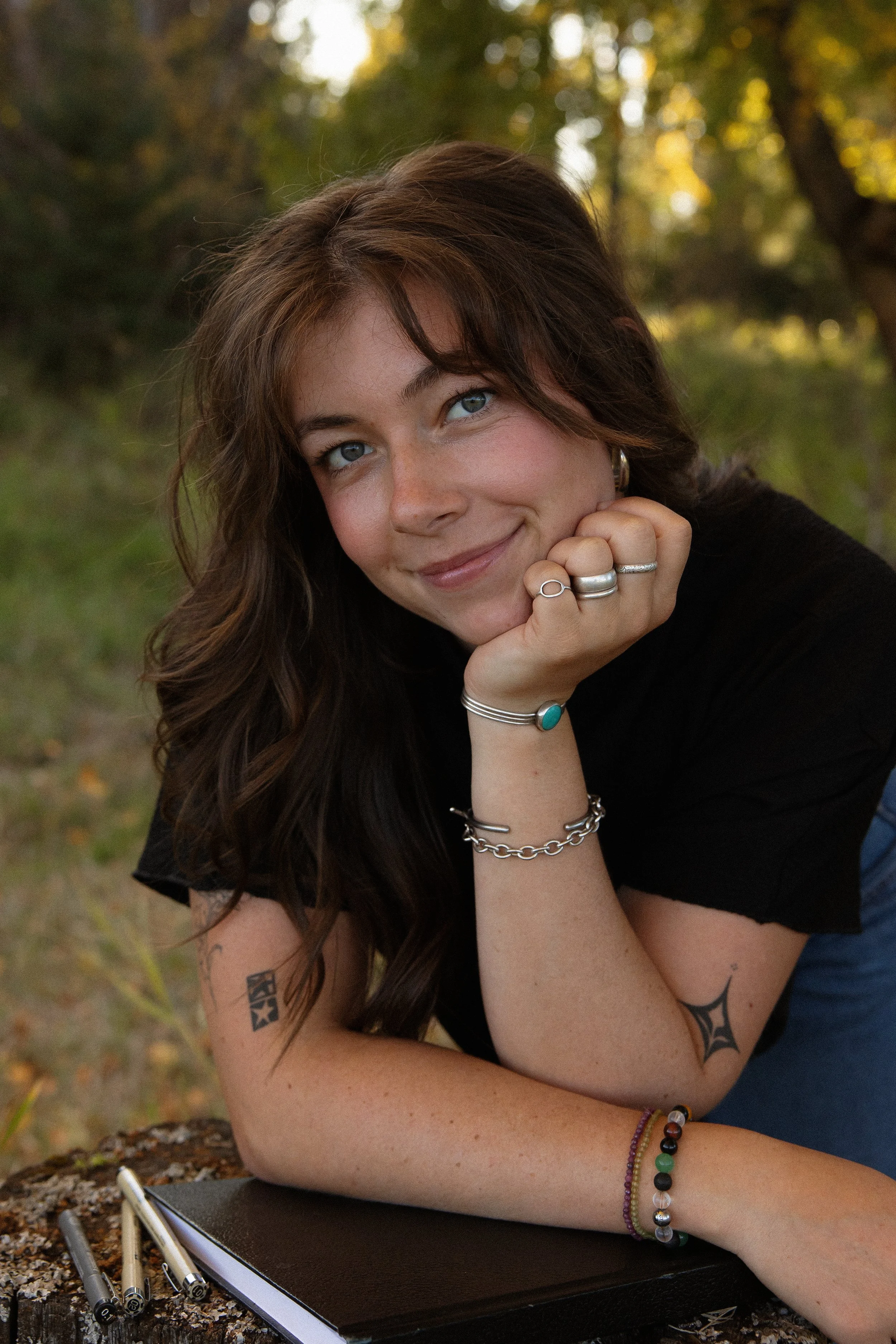 A young woman with long brown hair and blue eyes, wearing multiple rings and bracelets, rests her chin on her hand and smiles at the camera outdoors in a wooded area with dappled sunlight.