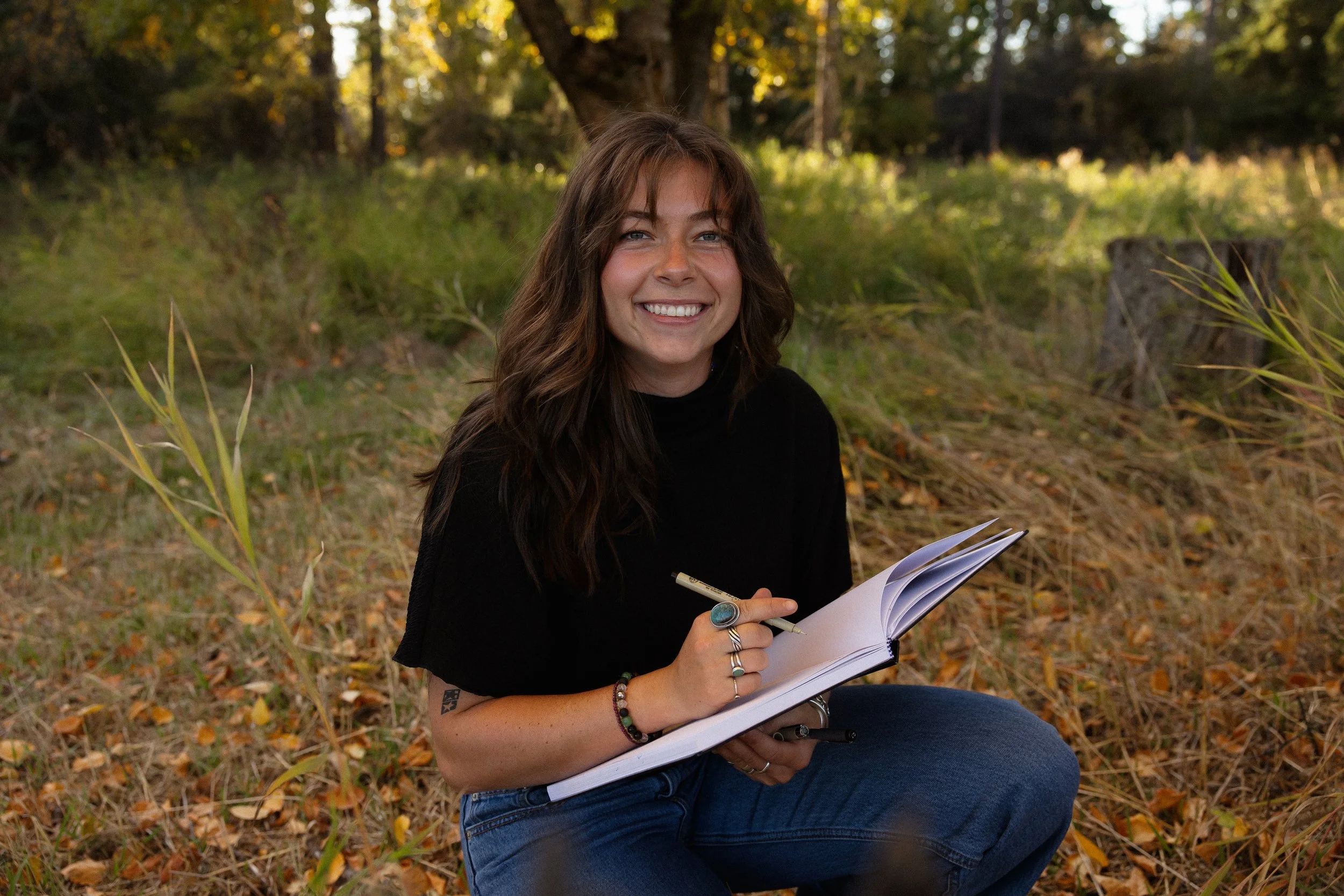 A woman with long, dark hair and a broad smile is sitting outdoors under a tree, holding a notebook, a pen, and a pencil in her hands. She is wearing a black shirt and blue jeans, and has several rings and bracelets. The background features a lush, green area with trees and fallen leaves.