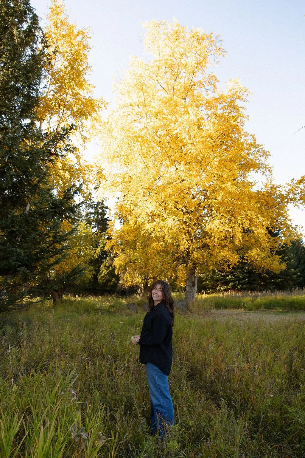 A woman standing in a grassy field during fall, with yellow trees and a clear sky in the background.