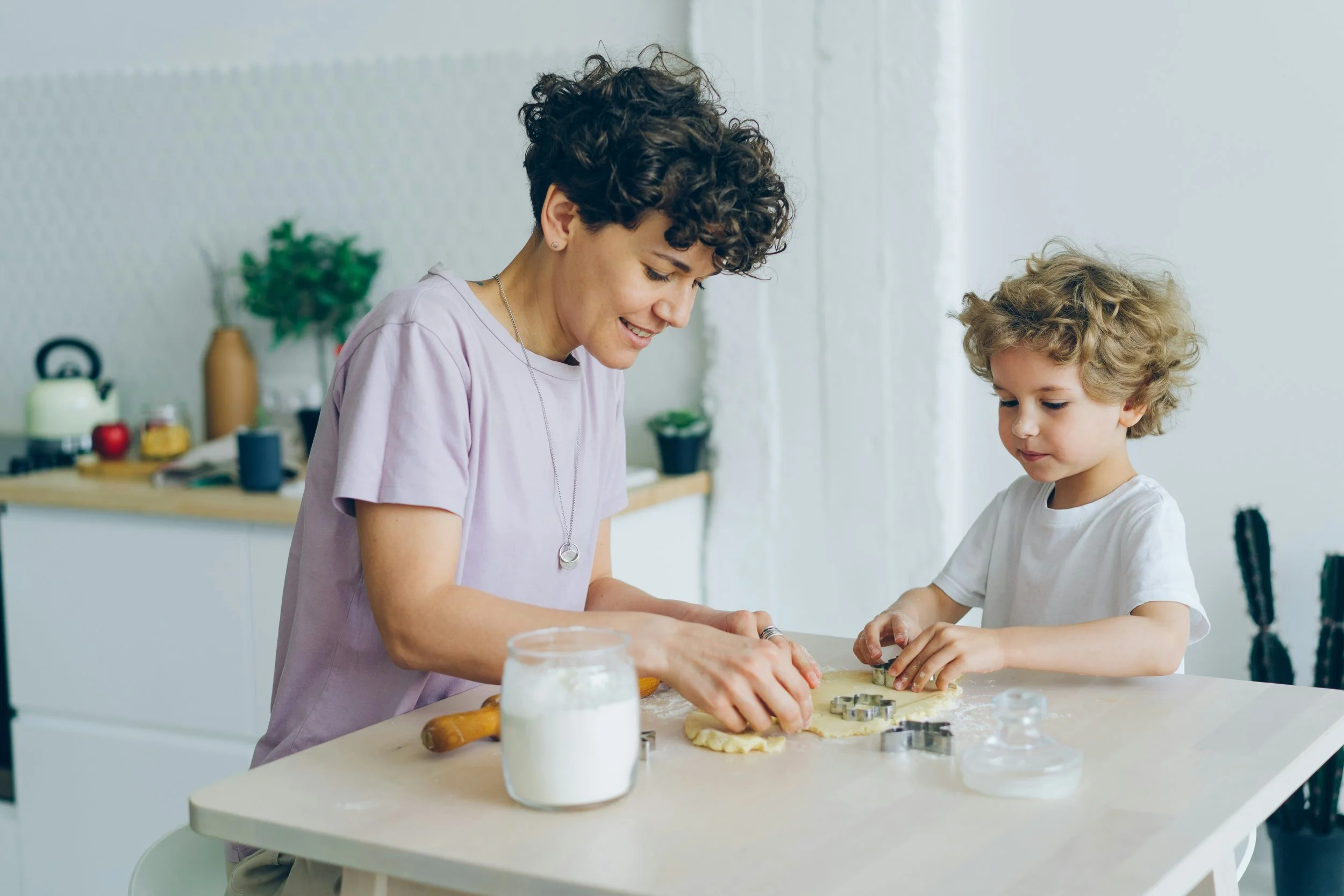 A woman and a young boy baking cookies together in a bright kitchen. They are using cookie cutters on rolled dough, with baking ingredients on the counter.