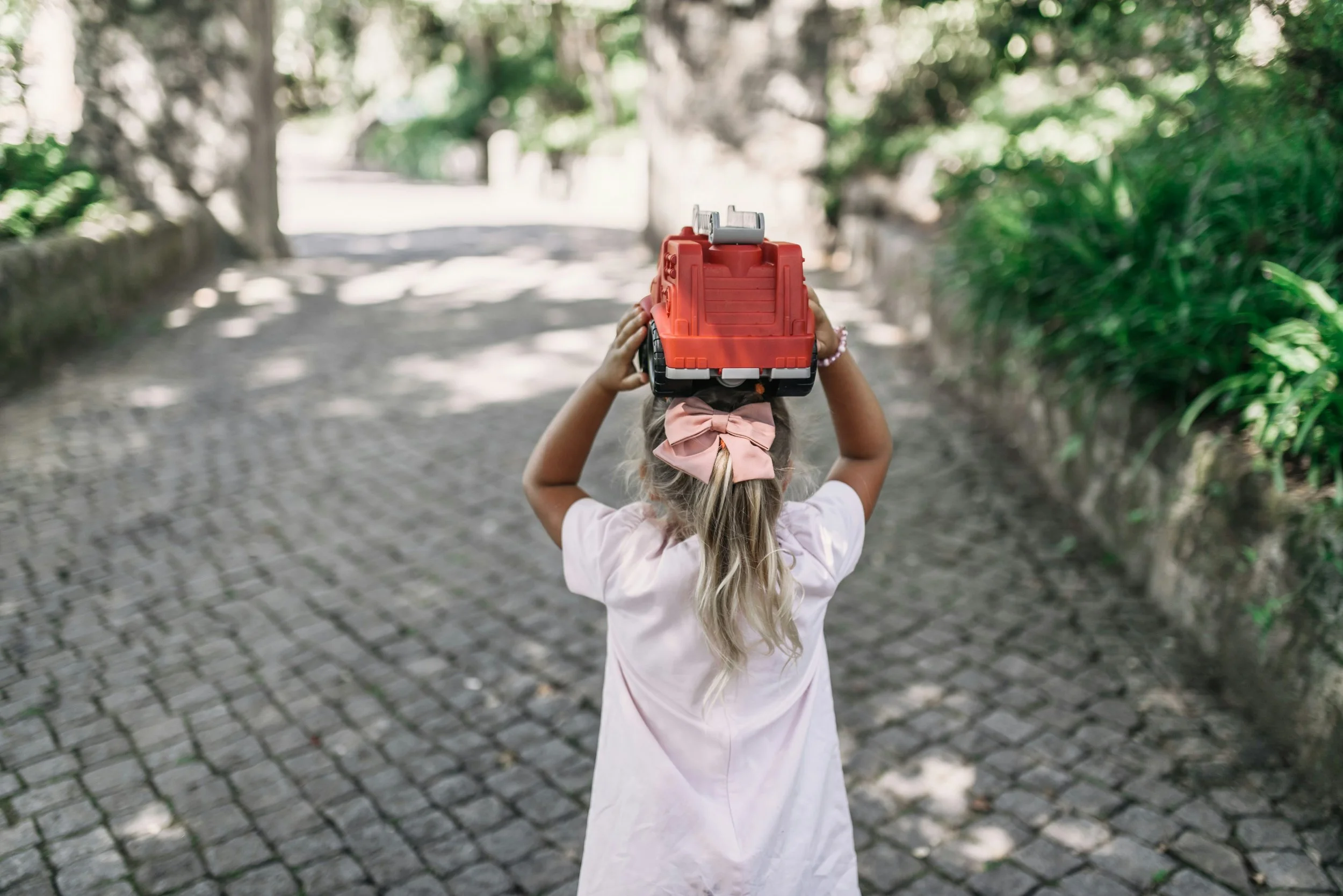A young girl with a pink bow in her hair holding a toy fire truck above her head while standing on a cobblestone path surrounded by greenery.