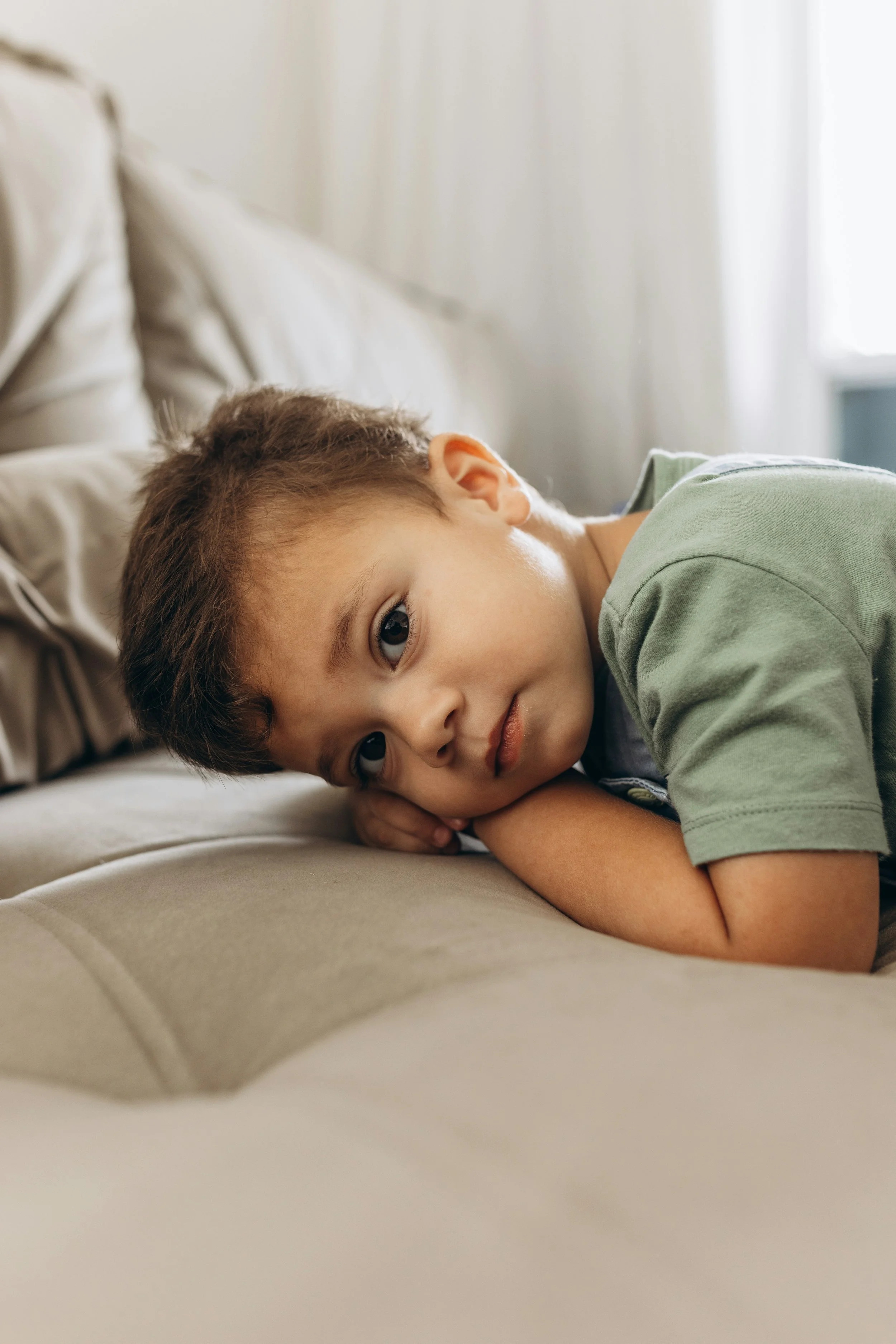 A young boy with short brown hair lying on his side on a bed, resting his head on his arms, looking at the camera with a calm expression.