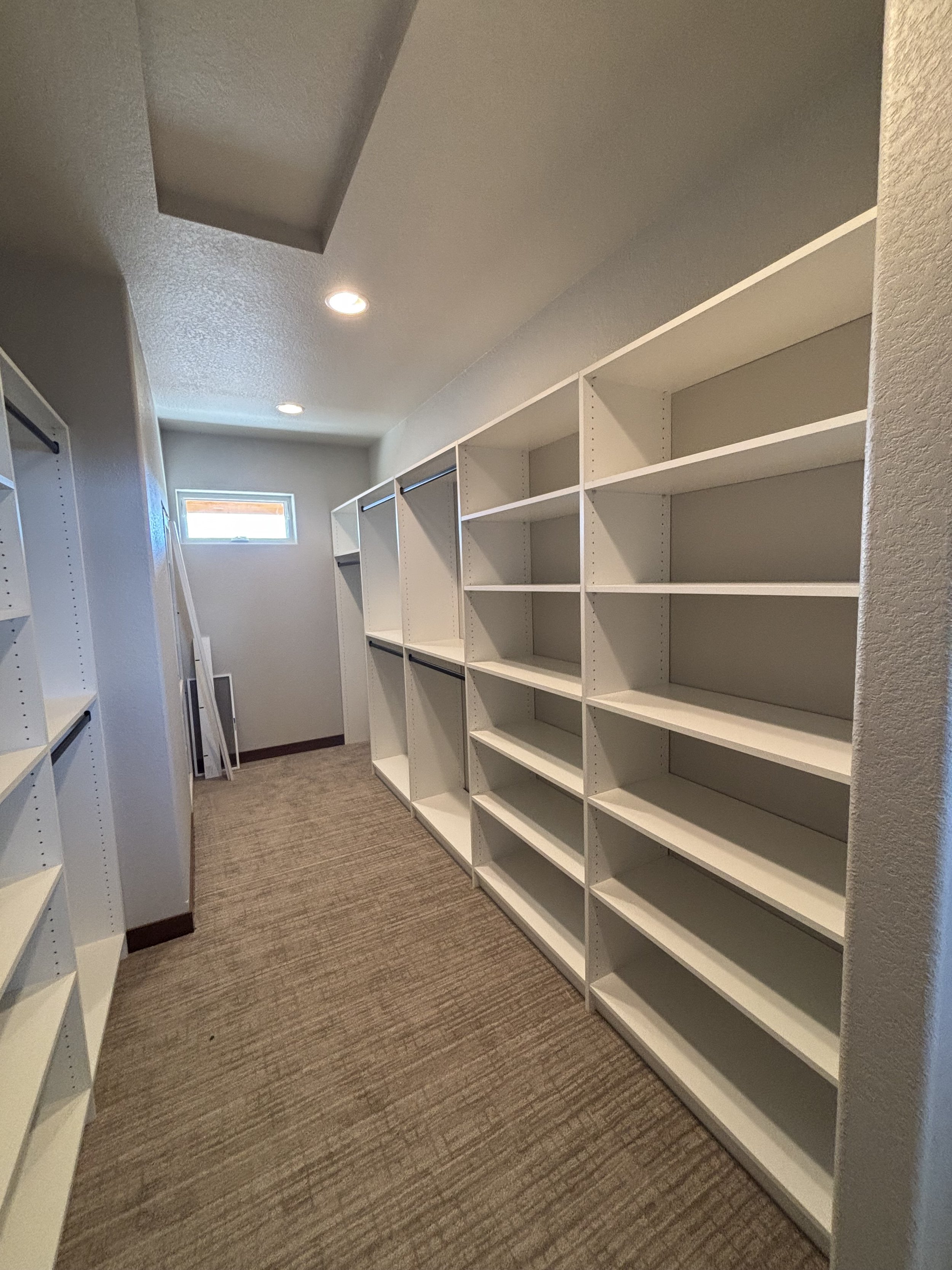 Empty white shelving units with multiple compartments in a small walk-in closet with beige carpet and a small window at the far end, ceiling with recessed lighting.
