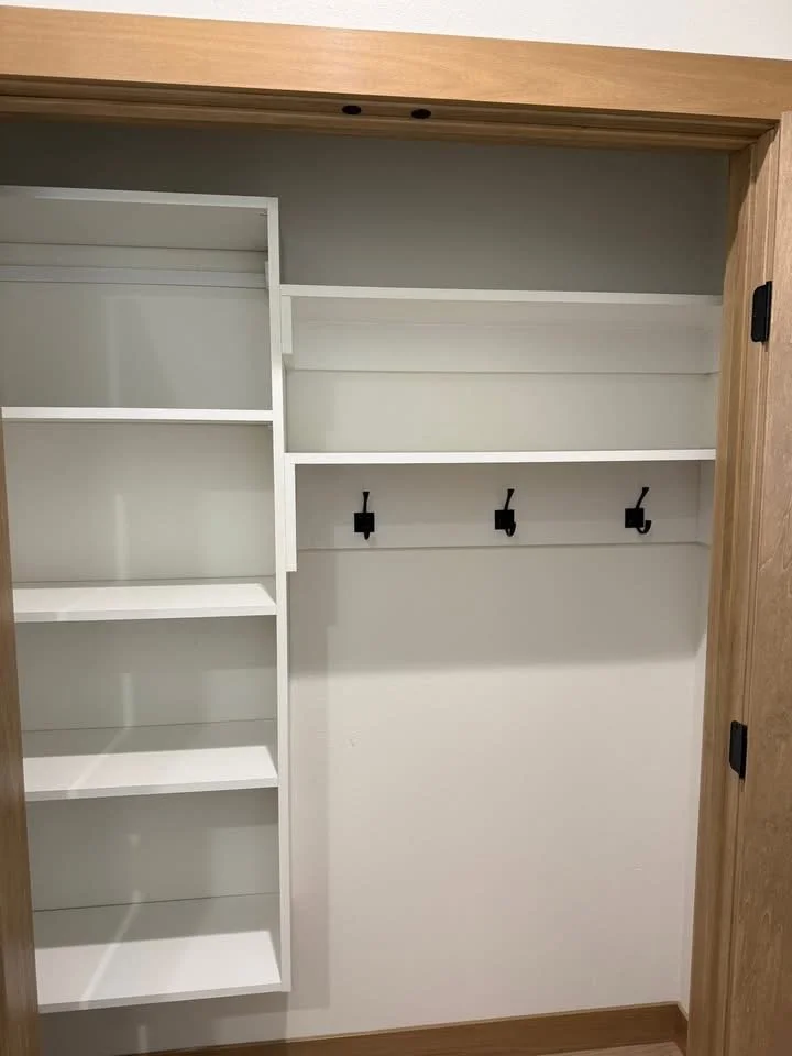 Empty closet with white shelves on the left, three hooks on the middle right wall, and a wooden door frame.