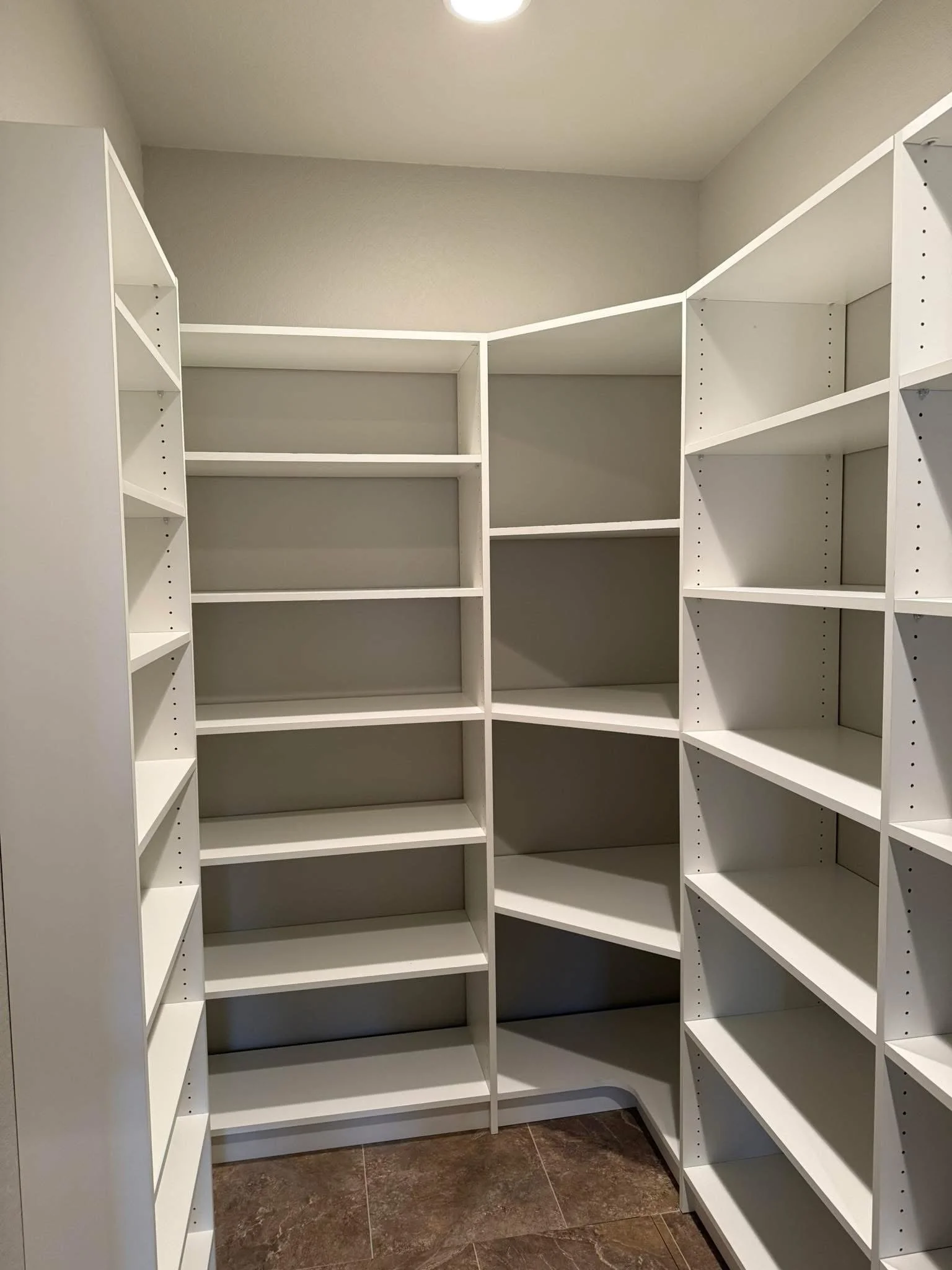 Empty white corner or L-shaped built-in shelving unit in a closet with multiple shelves, against gray walls, with a brown tiled floor.