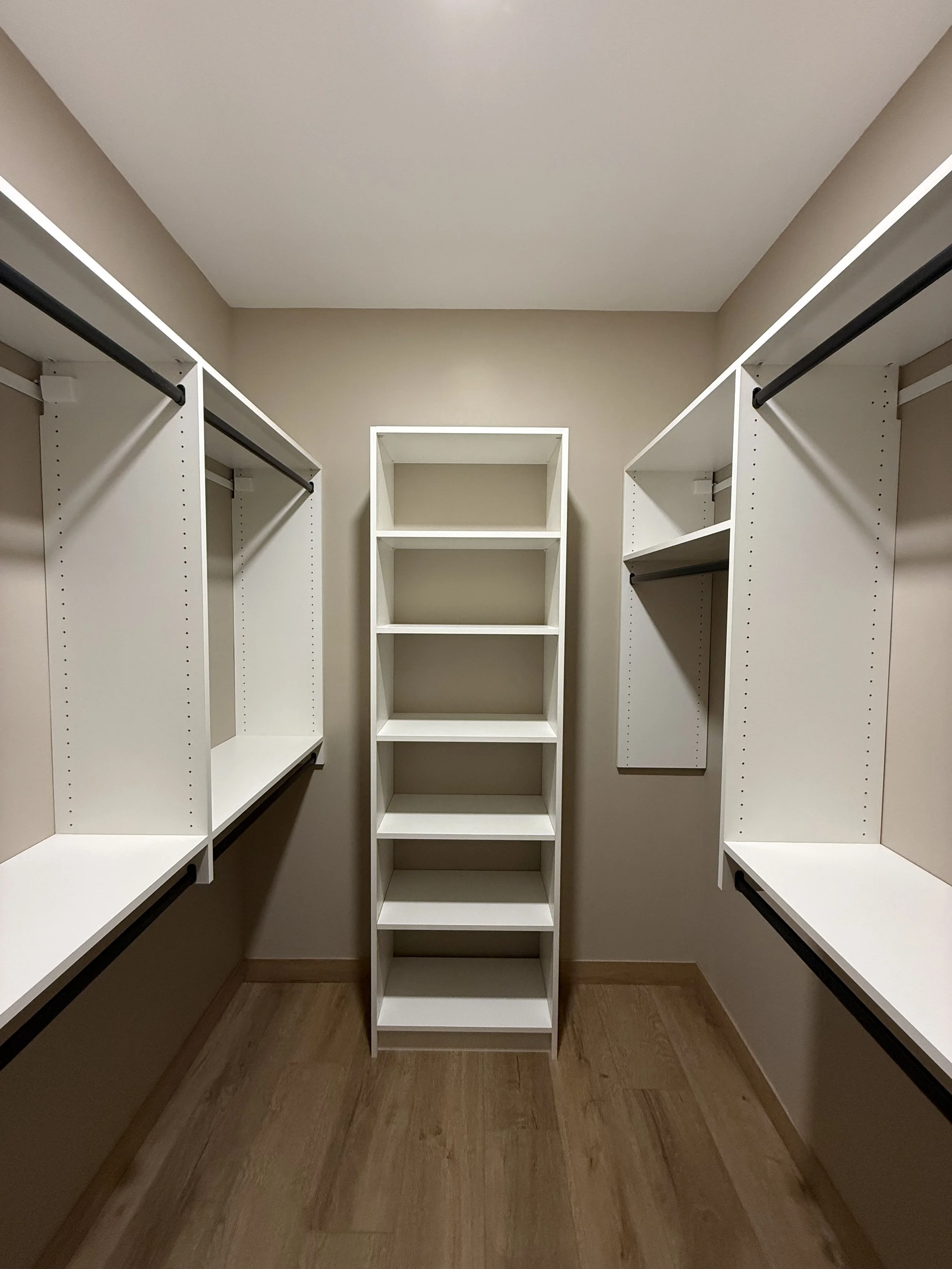 Empty walk-in closet with white shelving units, black rods for hanging clothes, and a beige wall and ceiling.