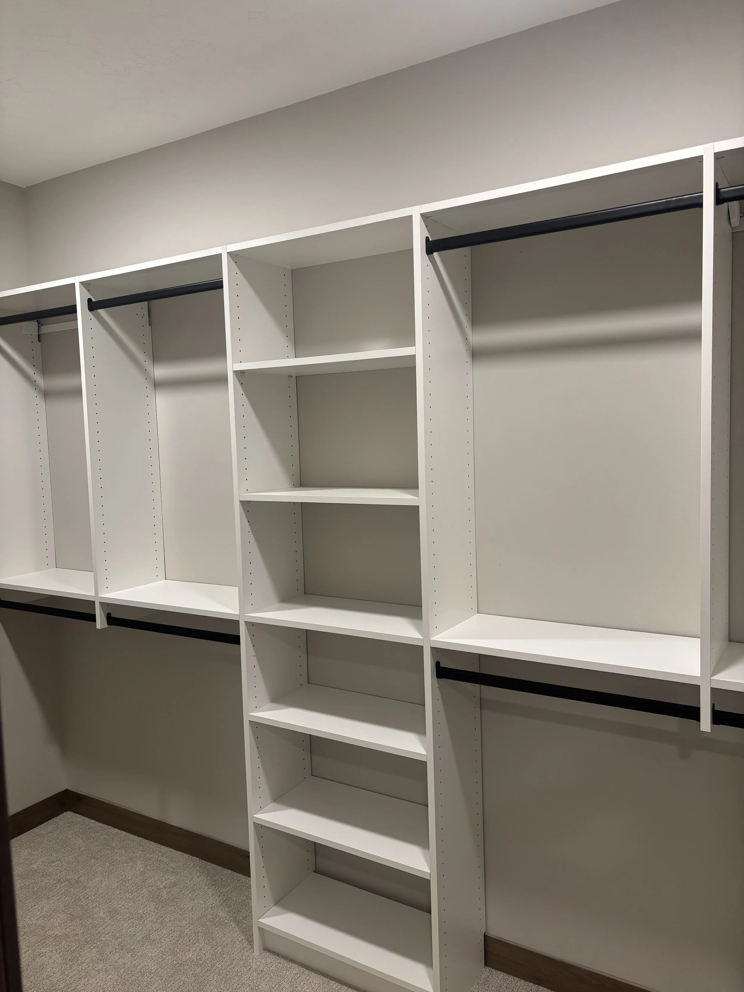 Empty walk-in closet with white shelves, black hanging rods, and beige carpeted floor.