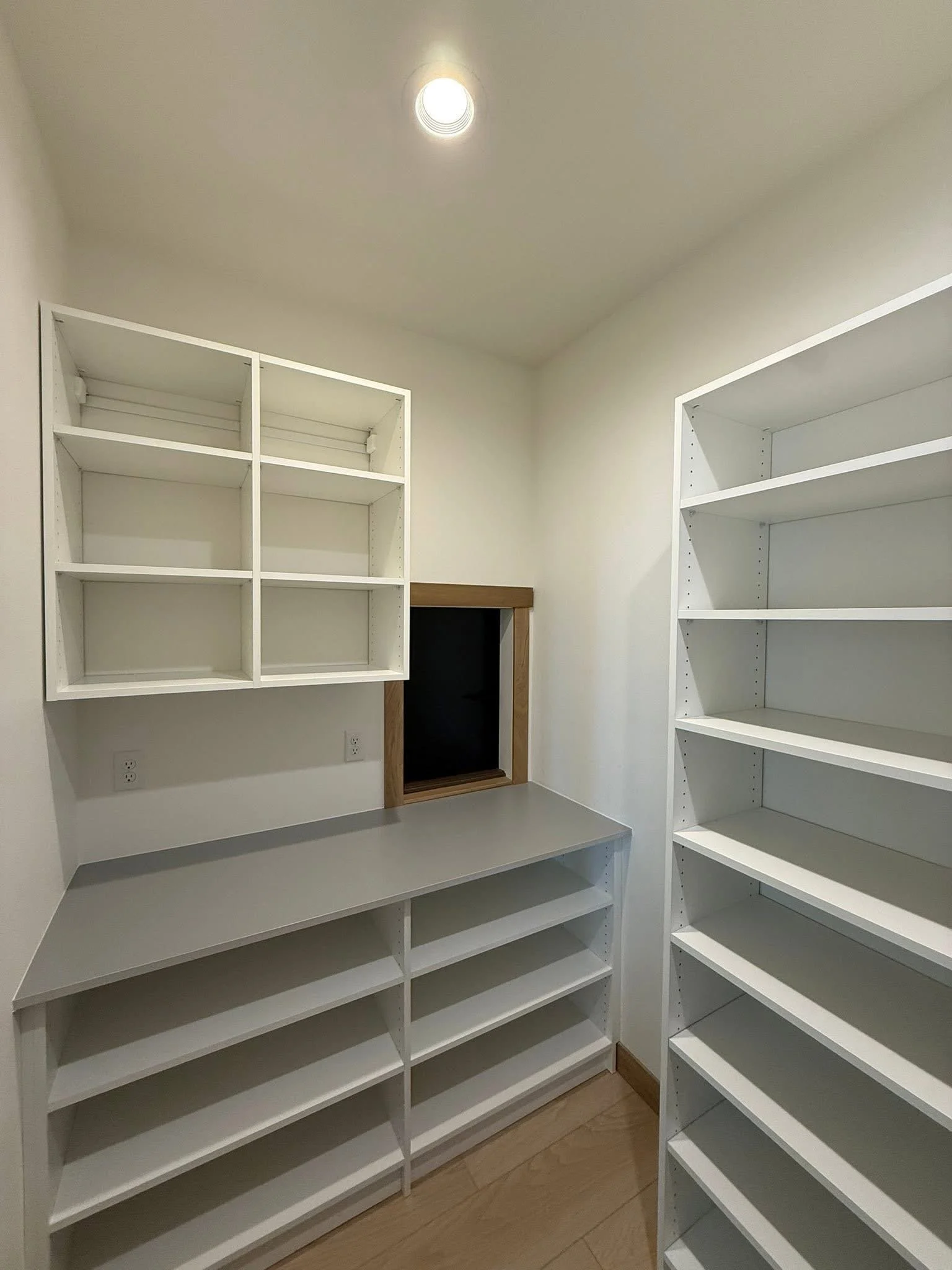 Empty white shelves and cabinets in a small room with a small window and a recessed light on the ceiling.