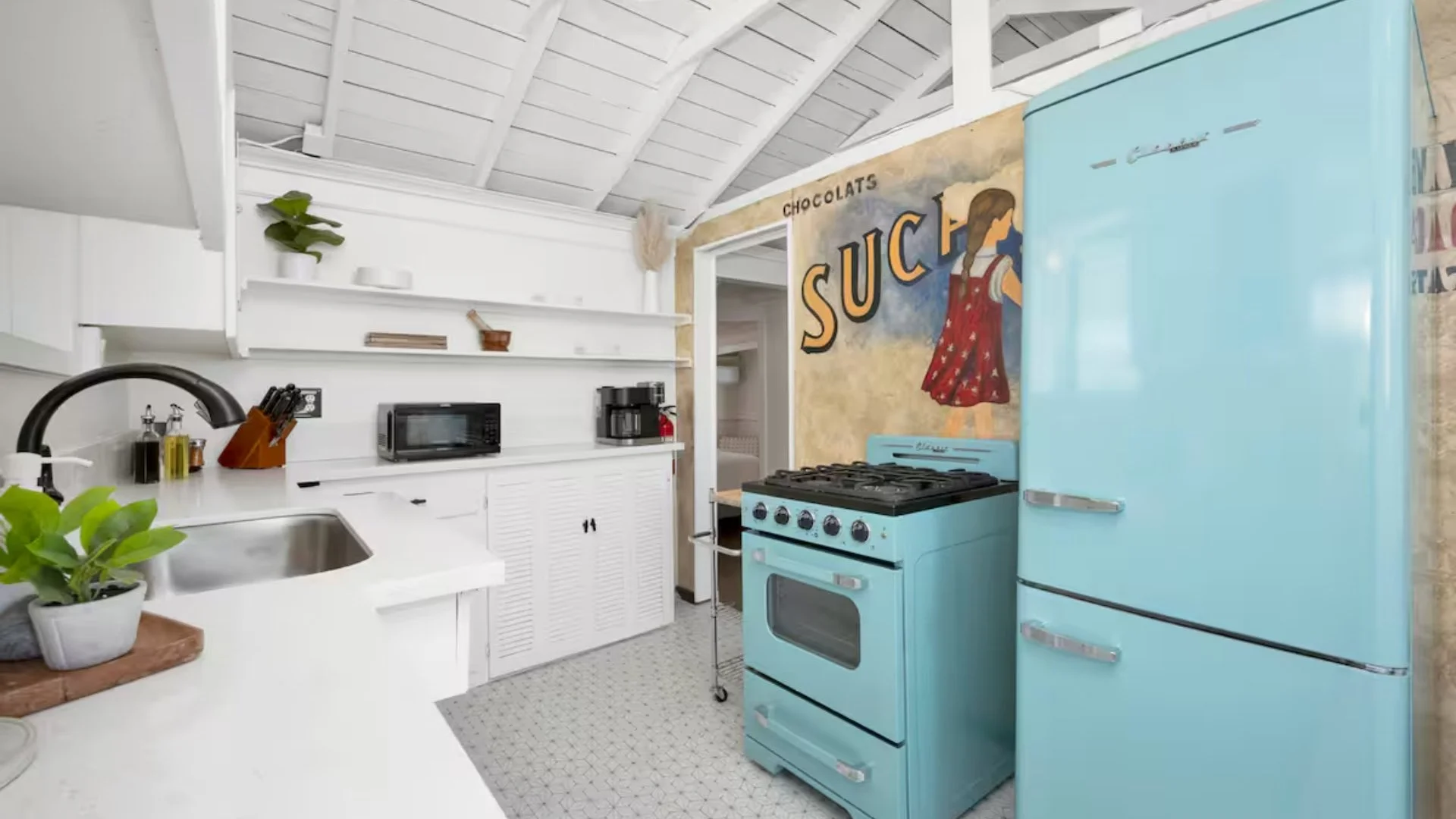 A kitchen with white cabinets, a black sink faucet, and open shelving on the wall with plants and dishes. A microwave and coffee maker on the counter. A teal retro refrigerator and stove near a wall with a mural that says 'Chocolats' and 'Suck' with 