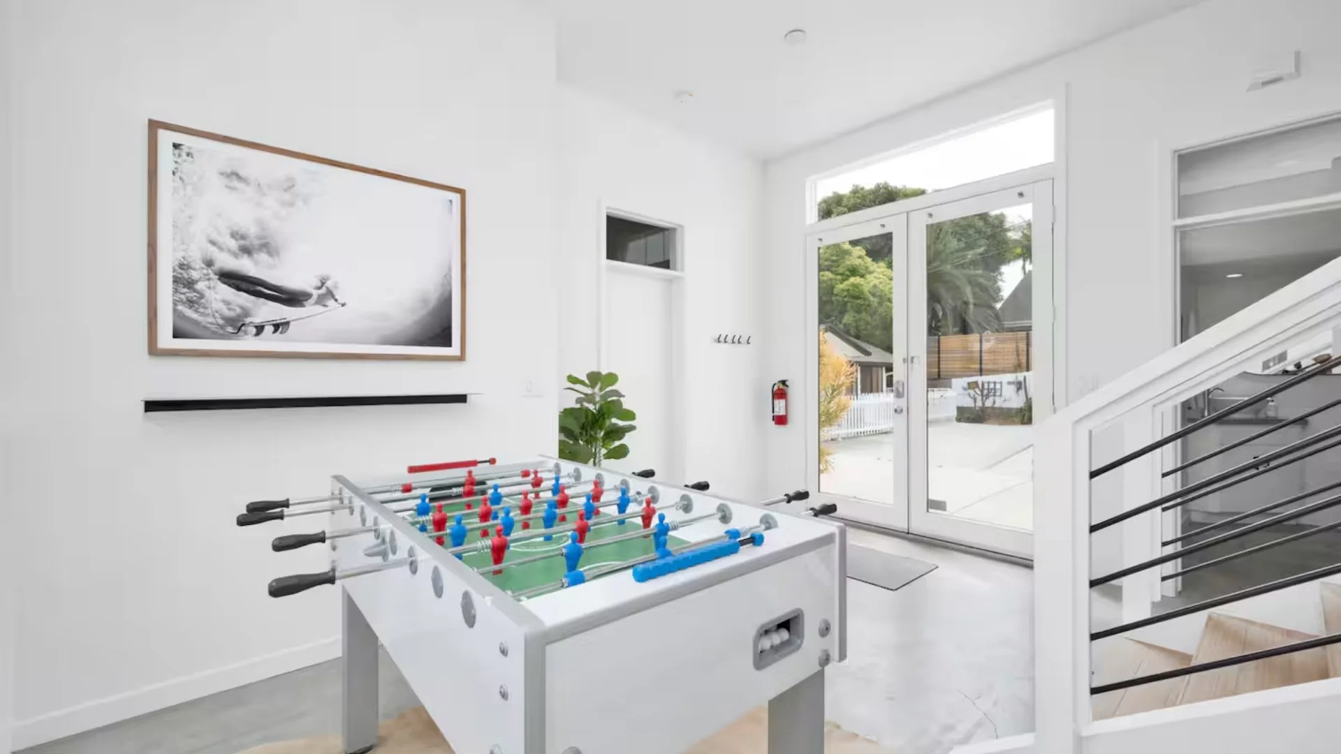 Indoor space with a foosball table, large glass front door, staircase with black handrails, framed black-and-white surfing photo, potted plant, coat hooks, and a fire extinguisher.