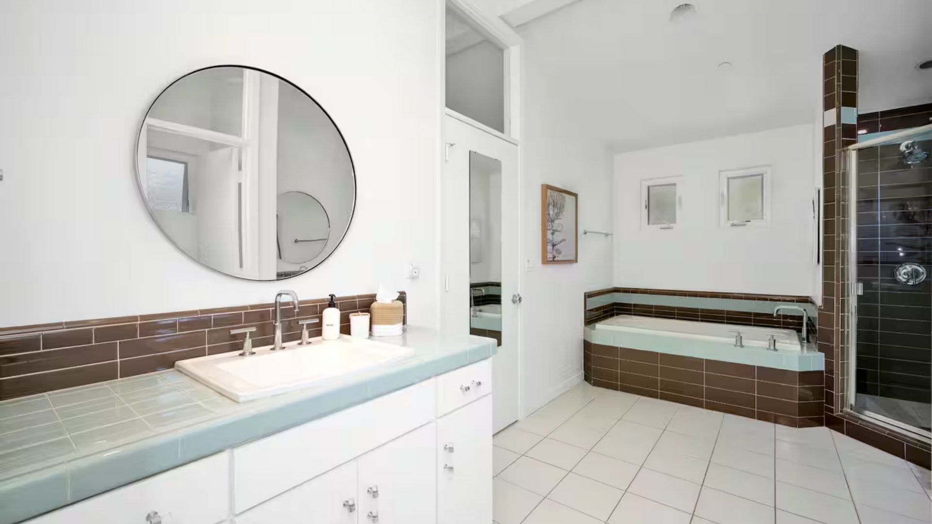 Bright bathroom with white walls, brown tile accents, a floating cabinet with a sink, and a large mirror. In the background, there's a bathtub with brown tile surround and a separate shower with black tile and glass door.