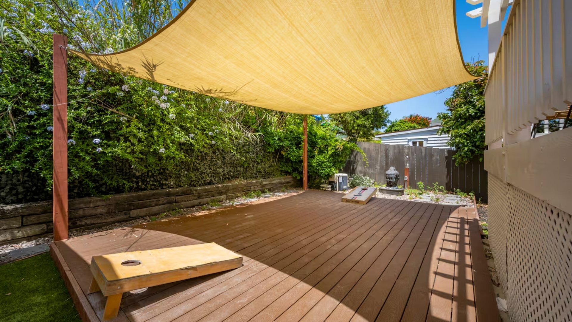 An outdoor wooden deck under a beige shade sail, with green bushes and trees behind a wooden retaining wall, and a fenced yard with decorative sculptures in the background.