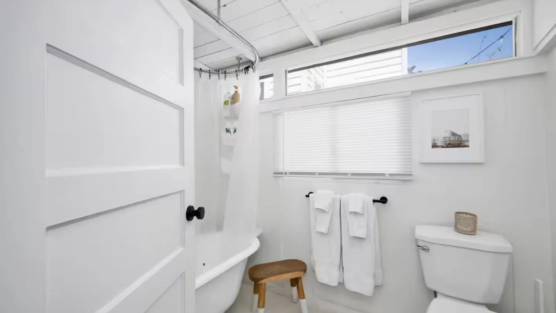 Bright white bathroom with a clawfoot bathtub, a small wooden stool, towels on a black rod, window blinds, framed photo of a vintage car, and a small basket on the toilet tank.