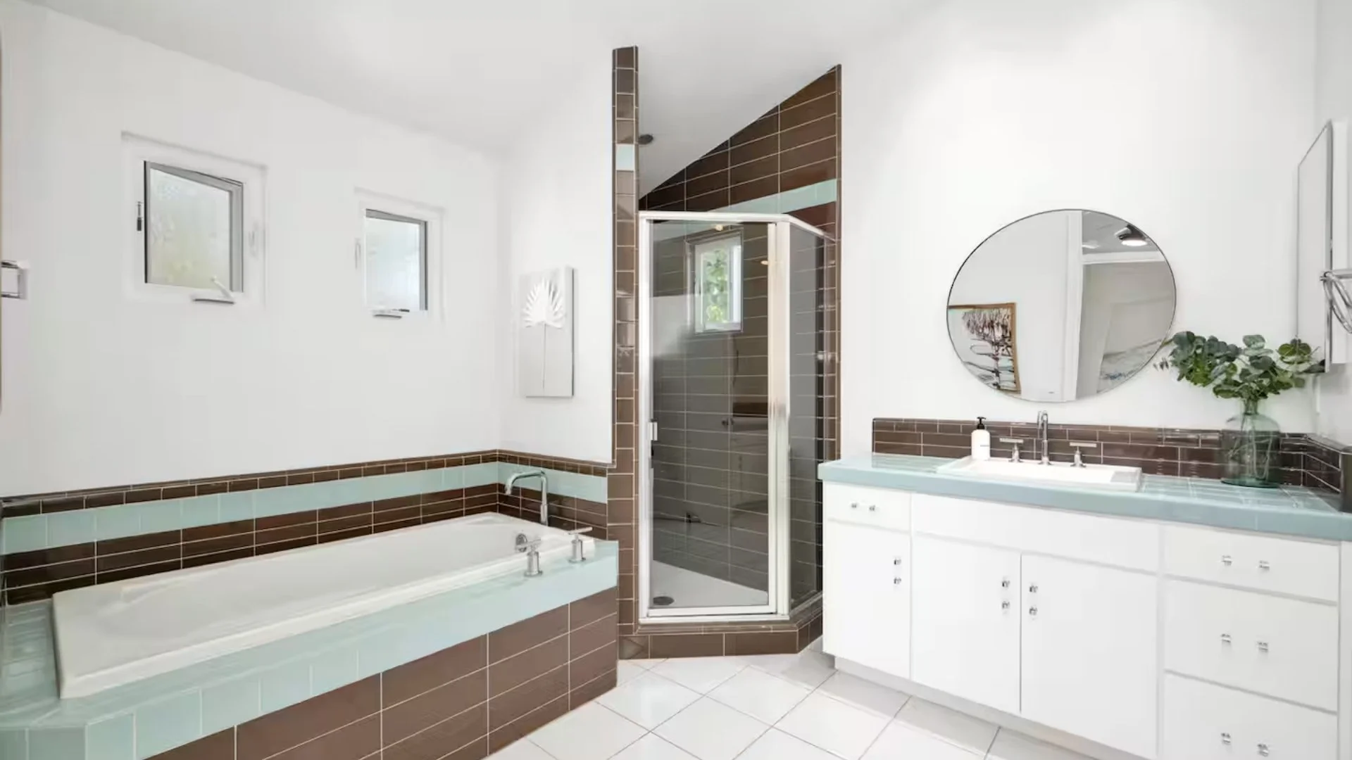 Modern bathroom with white walls, a large bathtub, a glass-enclosed shower, a white vanity with a round mirror, and decorative brown and blue tiles.