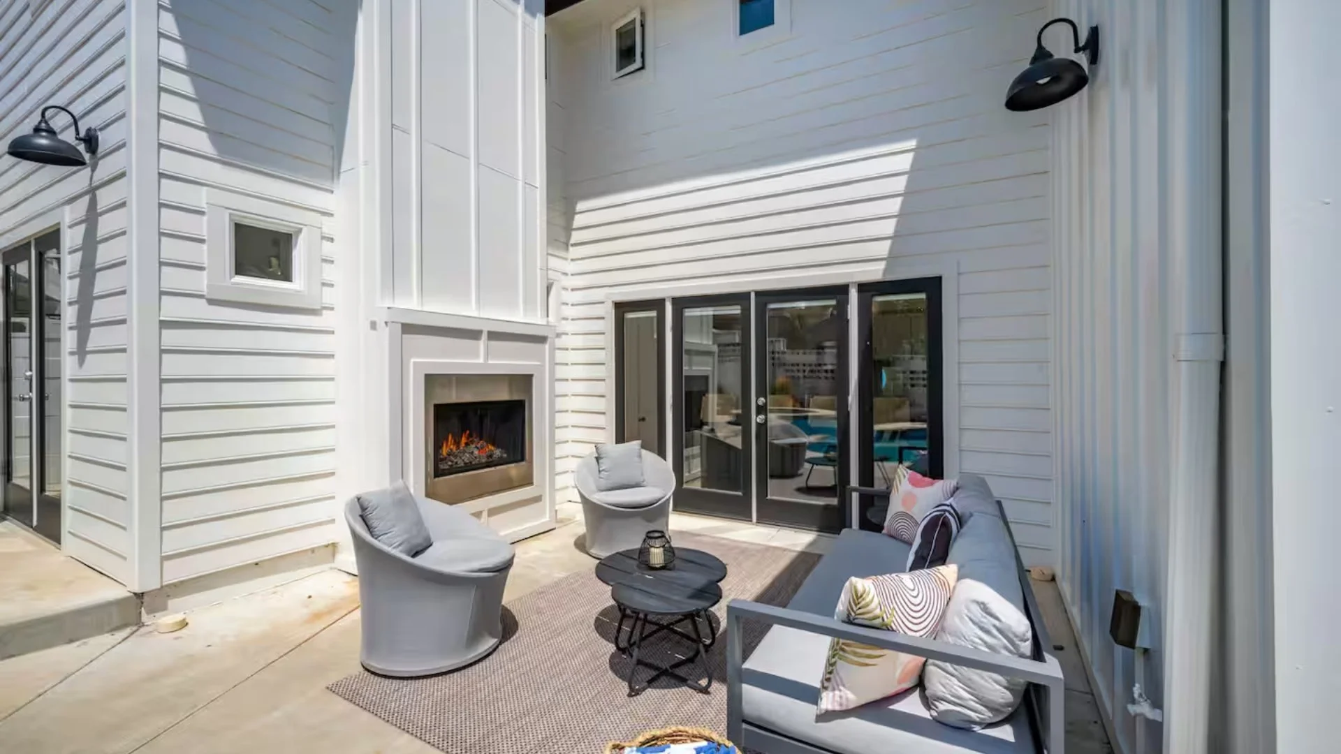 Outdoor patio area with white siding, black-framed glass doors, patio furniture including a sofa and chairs with cushions, a small black table, and a built-in fireplace on the house wall. Two black wall-mounted light fixtures are on either side of th