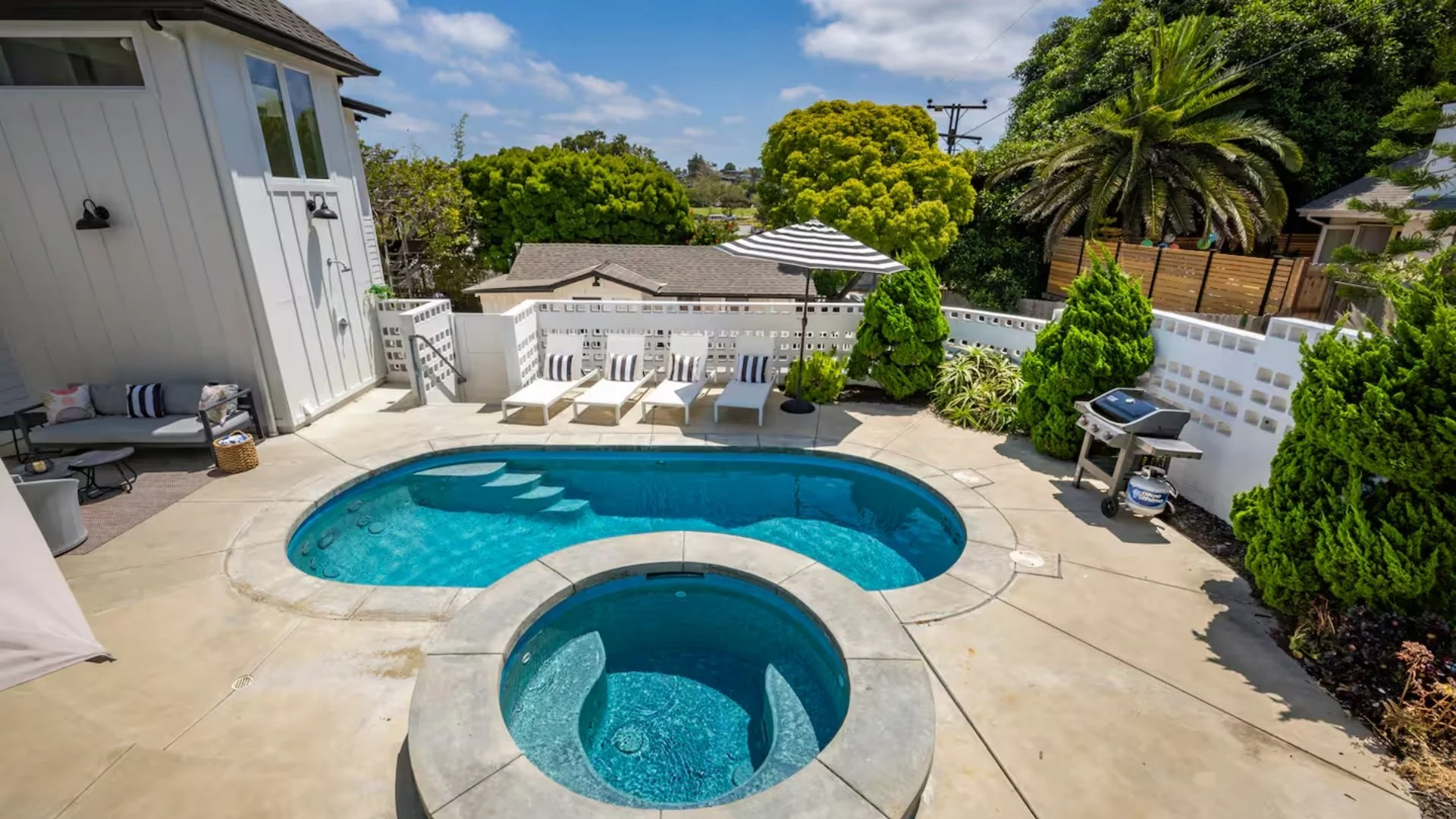 Residential backyard with a swimming pool and hot tub, surrounded by trees and bushes, with lounge chairs and an umbrella, on a sunny day.