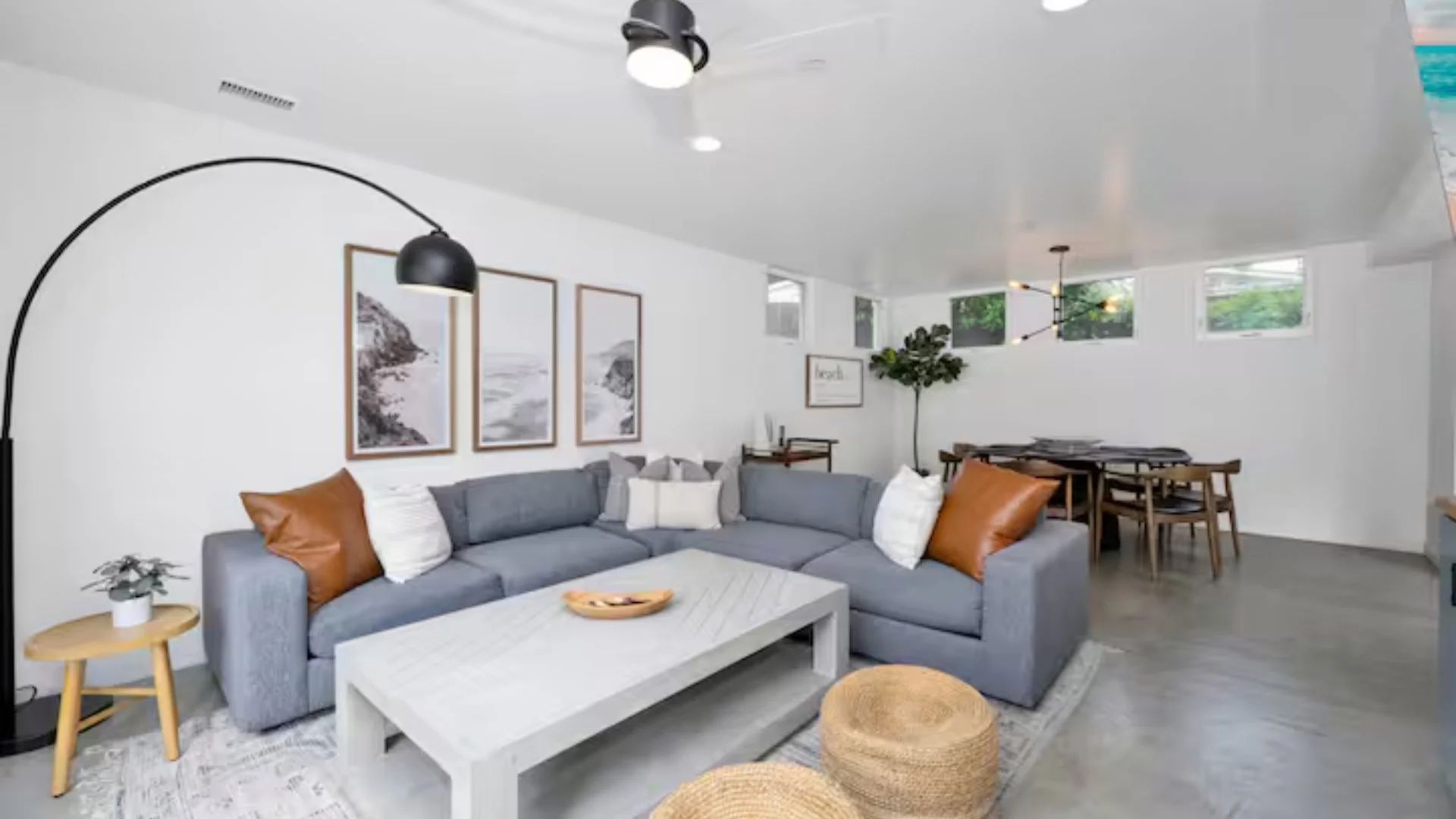 Living room with gray sectional sofa, white walls, framed black-and-white artwork, black arc floor lamp, white coffee table, small side table with plant, wicker poufs, dining area with wooden table and chairs, indoor plant, and modern lighting fixtur