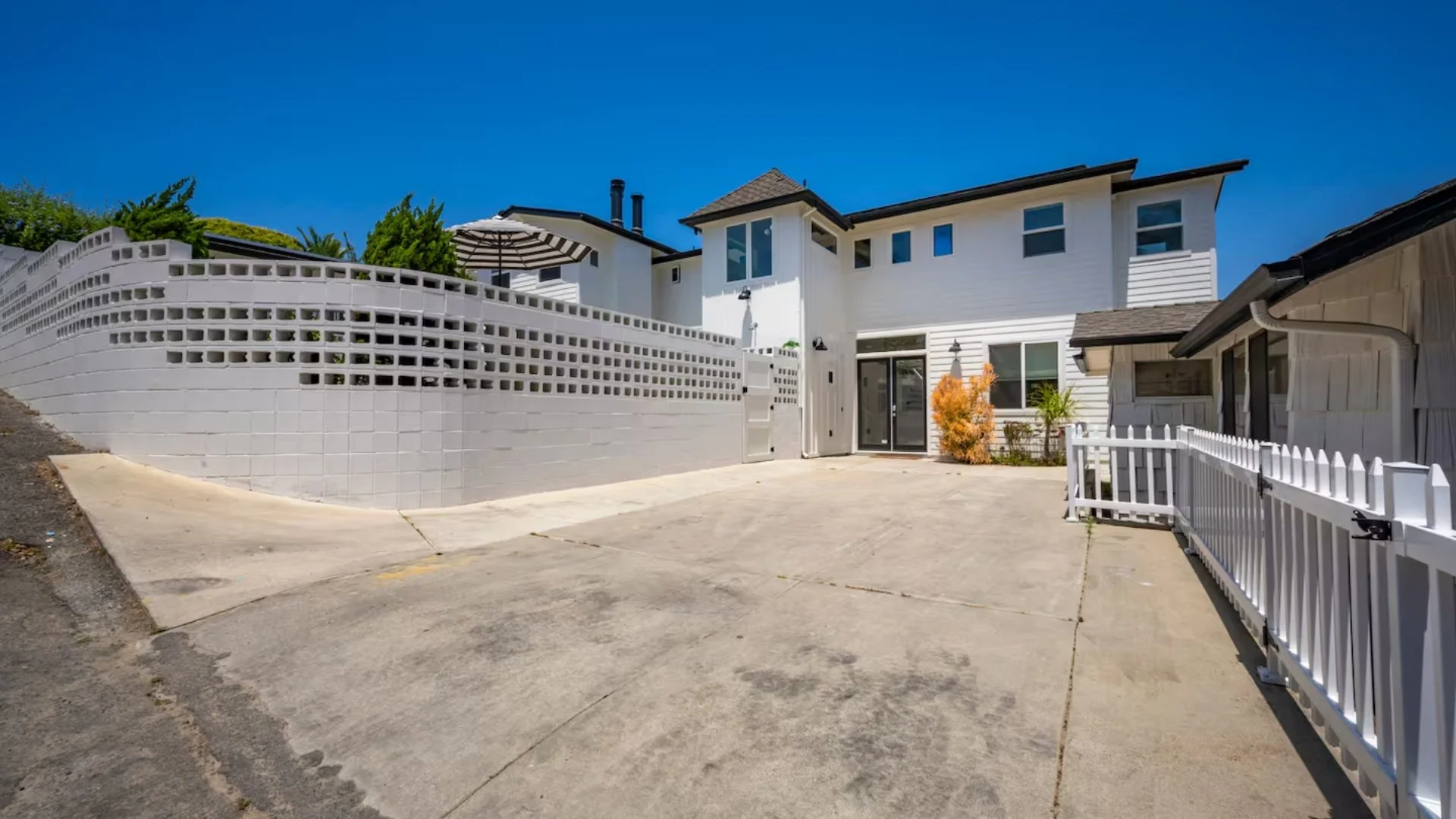 Backyard of a white house with a concrete driveway, a white fence, and a garden with plants, under a clear blue sky.