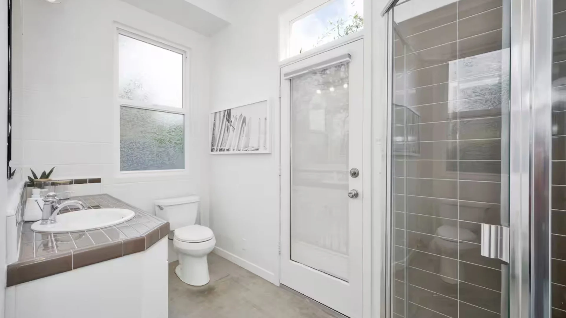 Small bathroom with white walls, a window, a toilet, a sink with a brown tiled countertop, and a glass shower door with brown tiles inside.