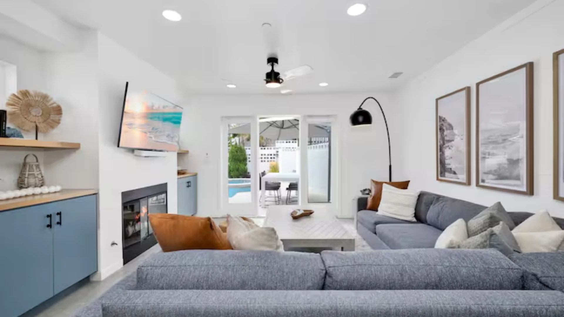 Living room with gray couches, a coffee table, wall-mounted TV, artwork on the wall, sliding glass door leading to a backyard with a pool, and various decorative items.