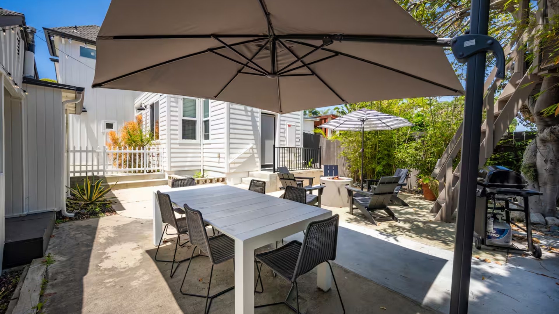 Backyard patio with outdoor dining table, chairs, umbrellas, lounge chairs, and a barbecue grill, surrounded by plants and a white house.