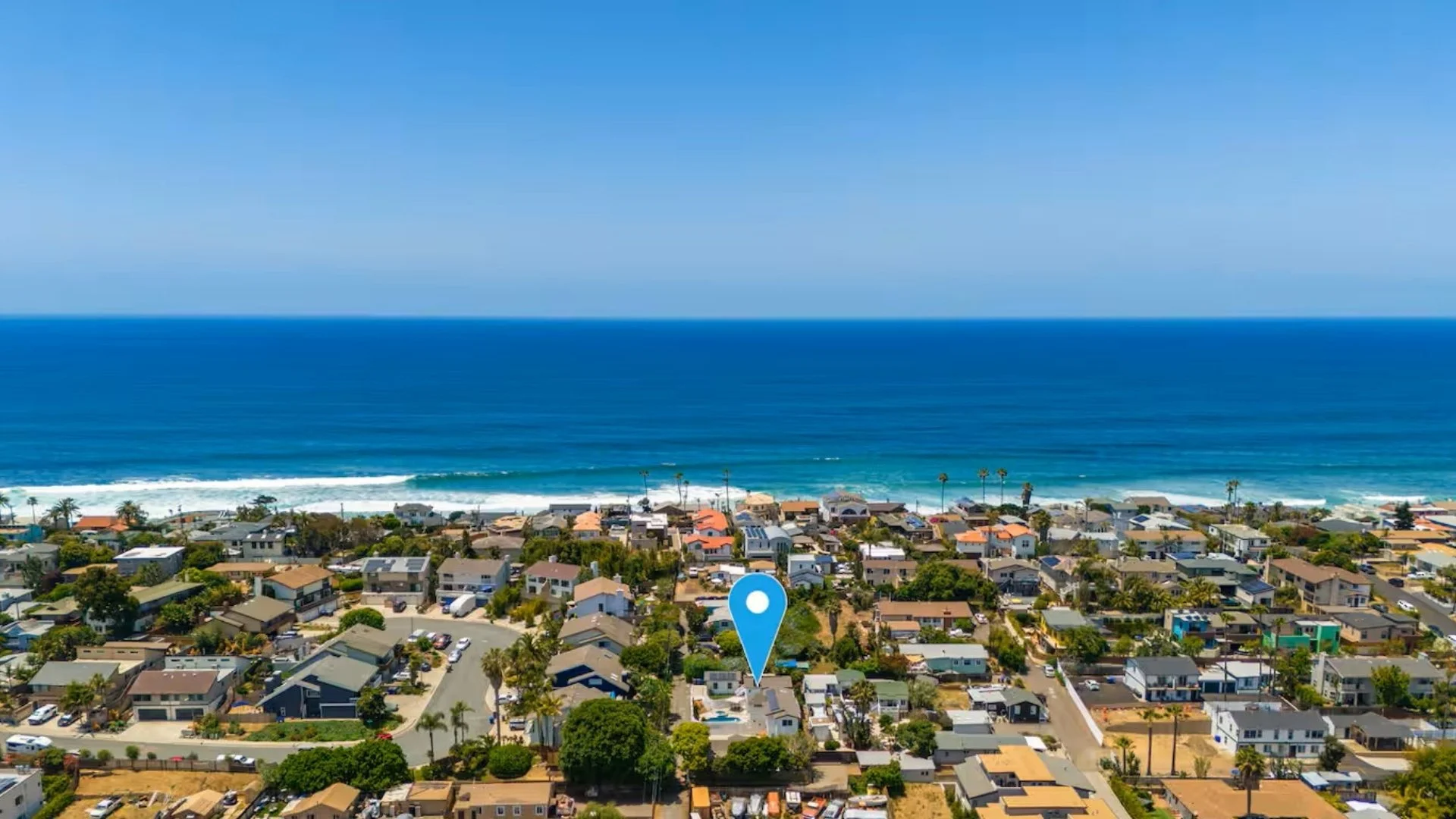 Aerial view of a beachside neighborhood with houses, trees, and streets near the ocean, with a blue map marker pointing to a specific house.