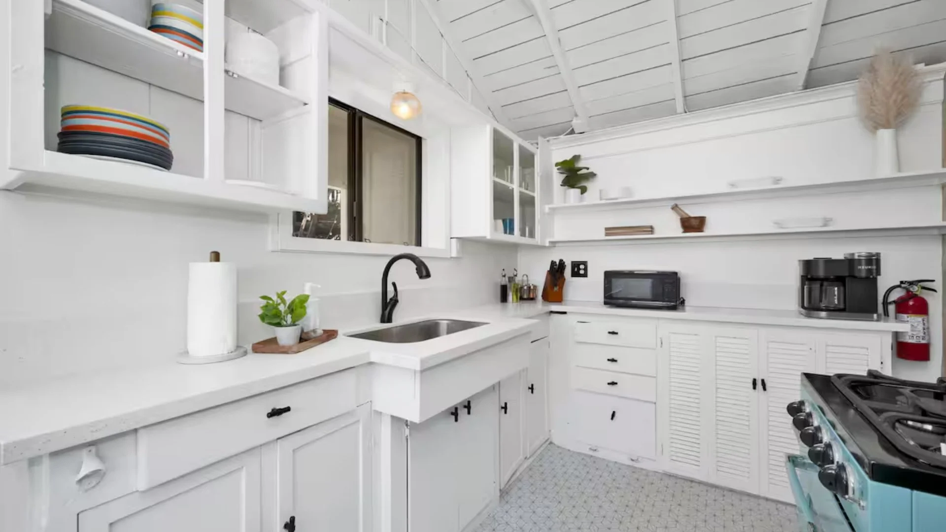 A bright, white kitchen with open shelving, a black faucet, small appliances, and decorative plants.