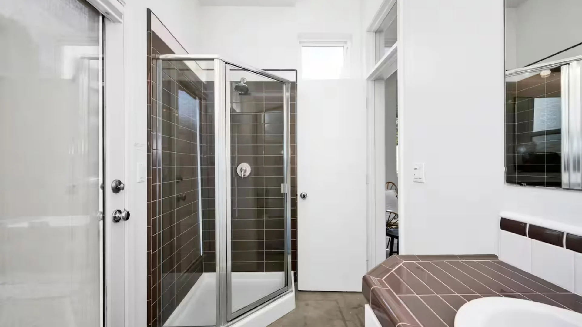 A bathroom with a brown-tiled walk-in shower, a brown-tiled countertop, and a white wall with a small window.