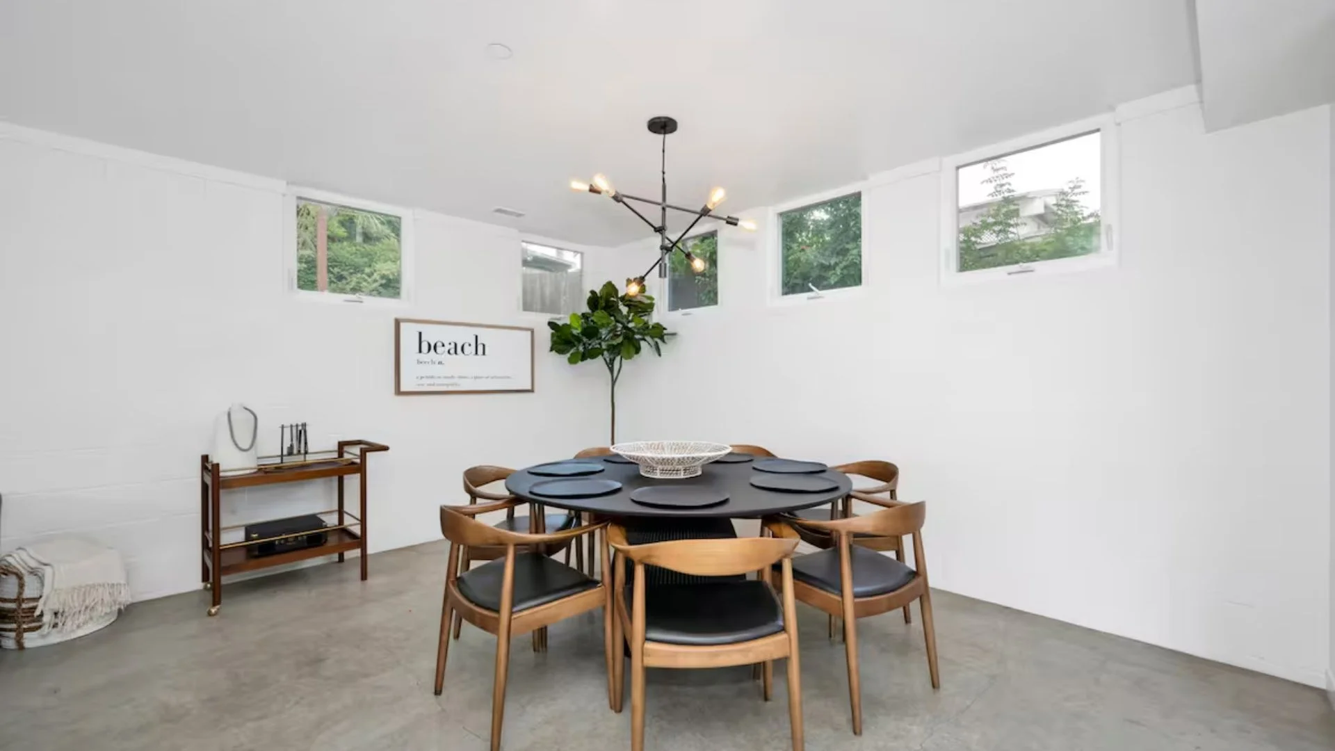 Dining room with a round table, six chairs, a modern chandelier, a wall art print that says 'beach,' and windows showing greenery outside.