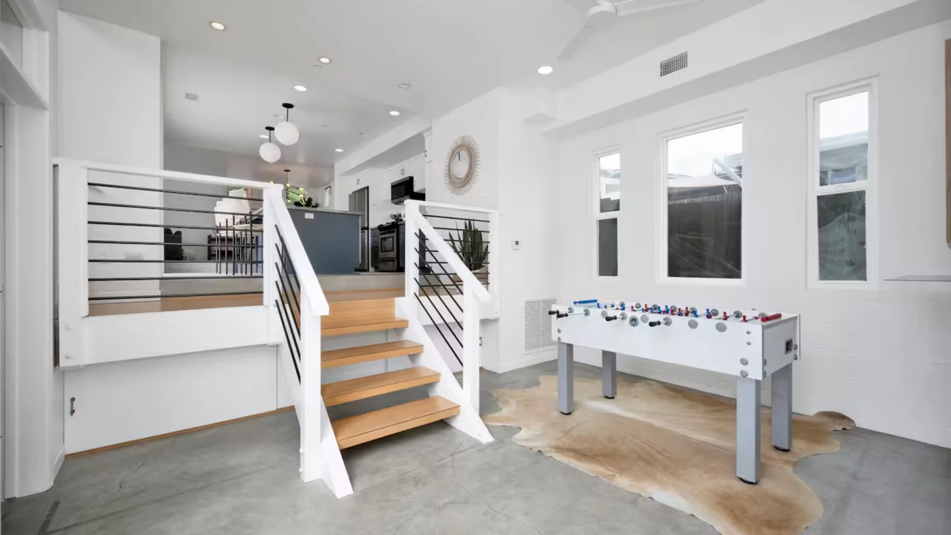 Bright modern living room area with a foosball table on a cowhide rug, staircase with wooden steps and black metal railings, white walls, large windows, and minimal decor.