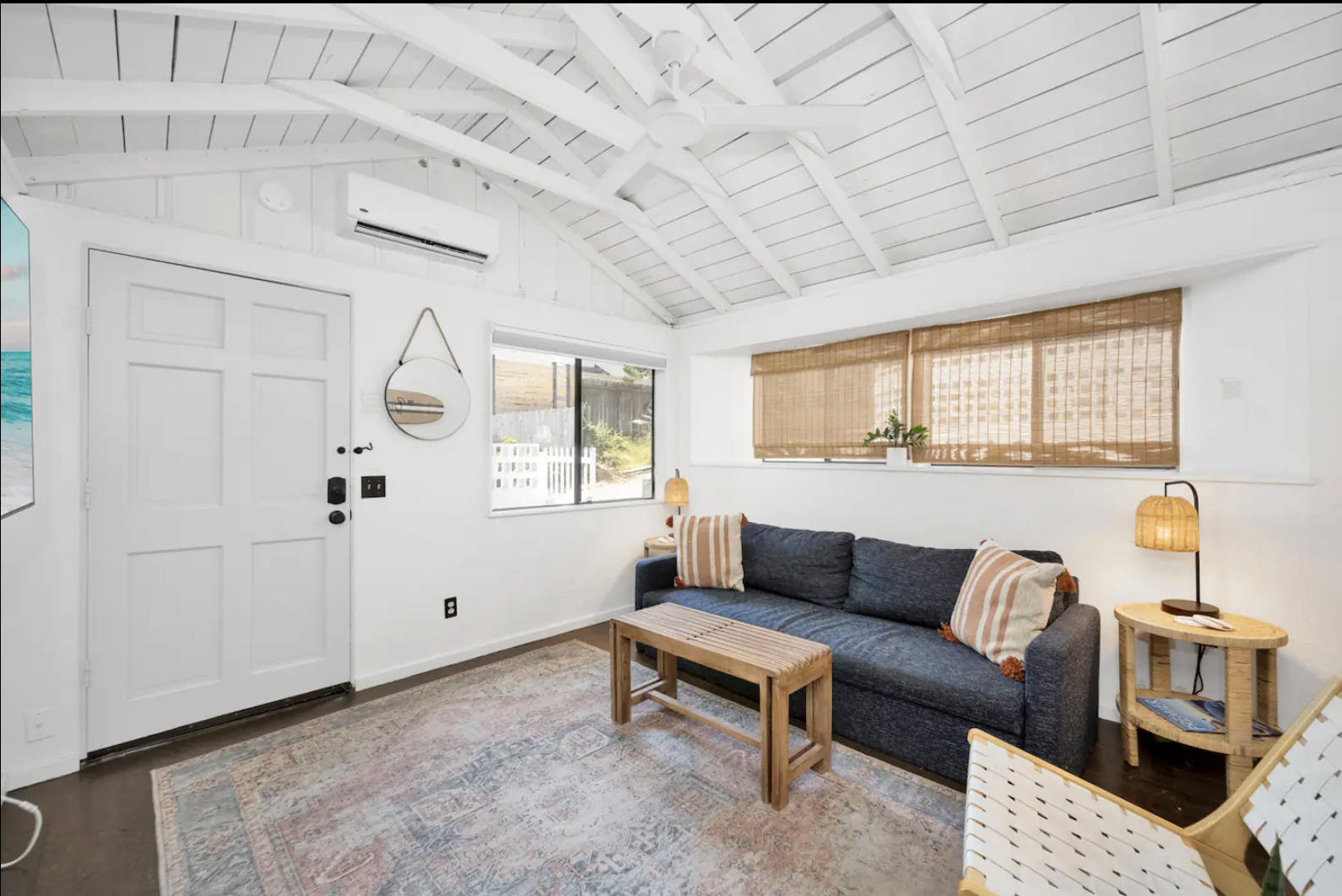 Living room with white walls, vaulted white ceiling, and window with bamboo blinds. Contains a dark blue sofa with striped pillows, a wooden coffee table, round side table, lamps, and a woven chair. There is a patterned area rug on the dark hardwood 