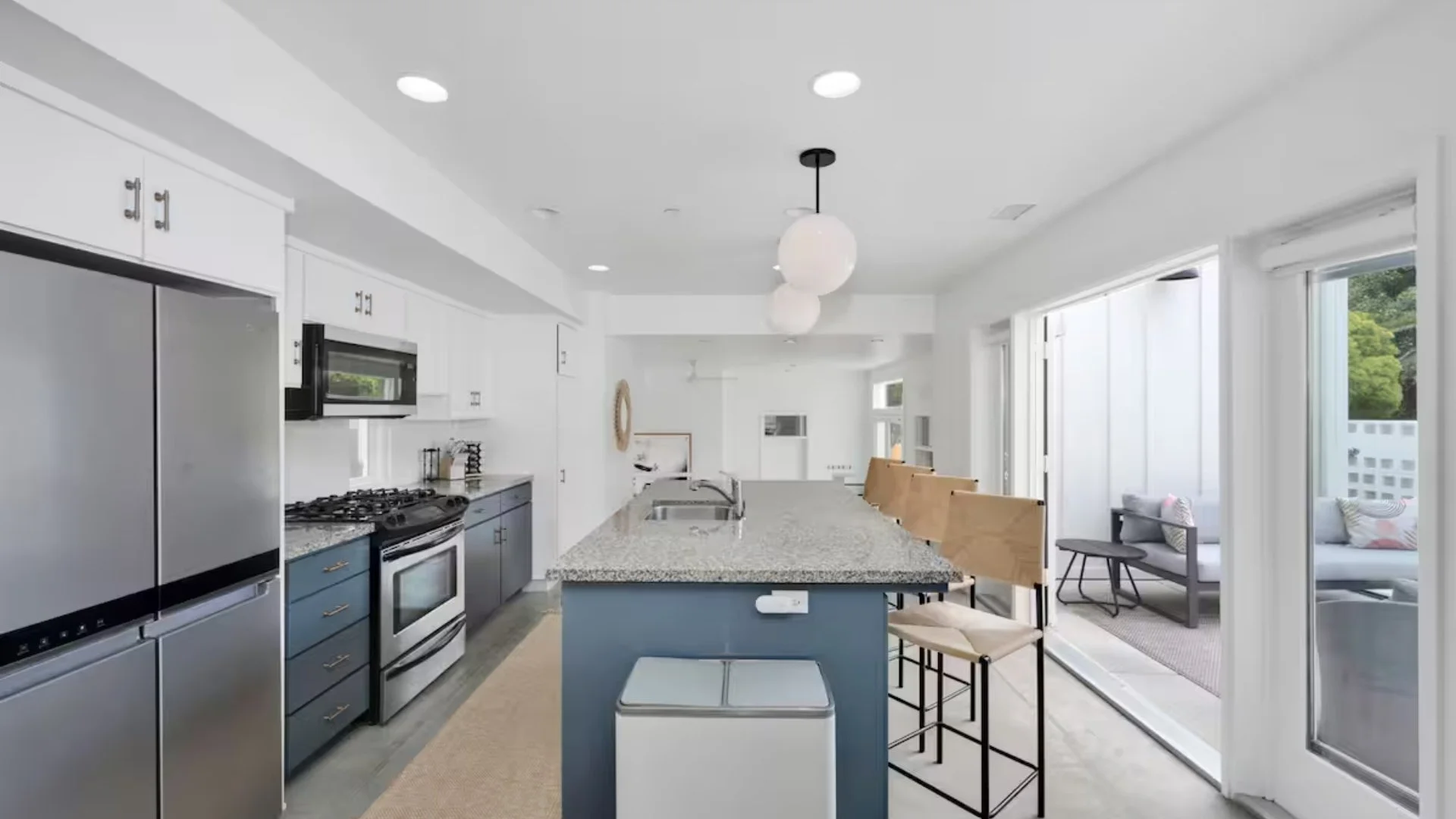 Modern kitchen with white cabinets, stainless steel refrigerator, granite countertops, island with seating, and adjacent sunroom with outdoor seating.