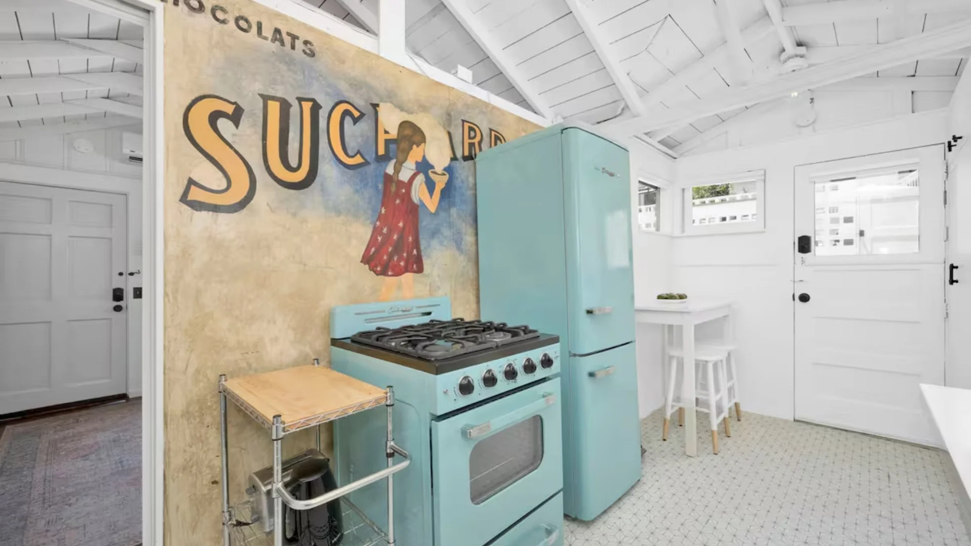 Bright white interior kitchen with vaulted ceiling, windows, light blue vintage refrigerator, gas stove, small table with two stools, and a mural with a girl drinking a beverage.