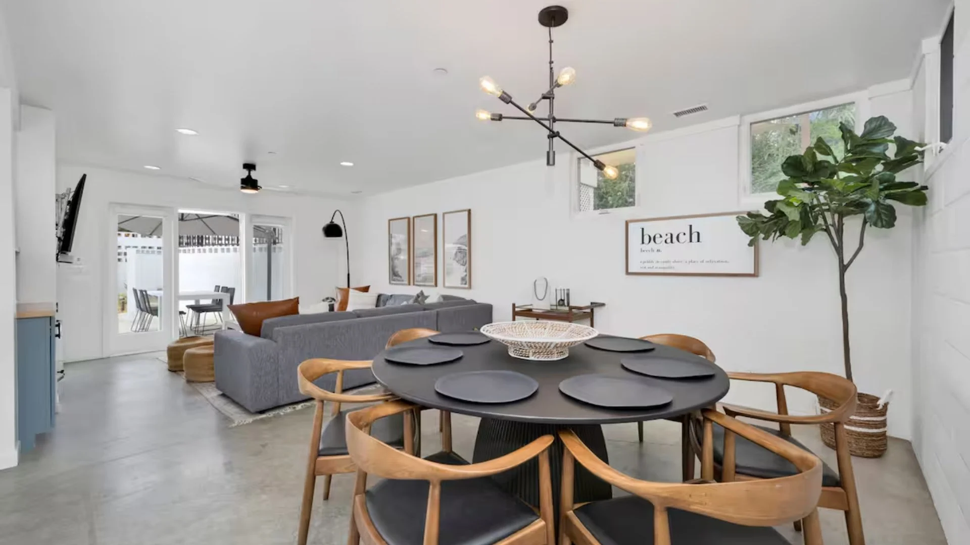Open-concept living and dining area with modern decor, light-colored walls, gray sectional sofa, wooden chairs around a black round dining table with black plates, and a large indoor plant.