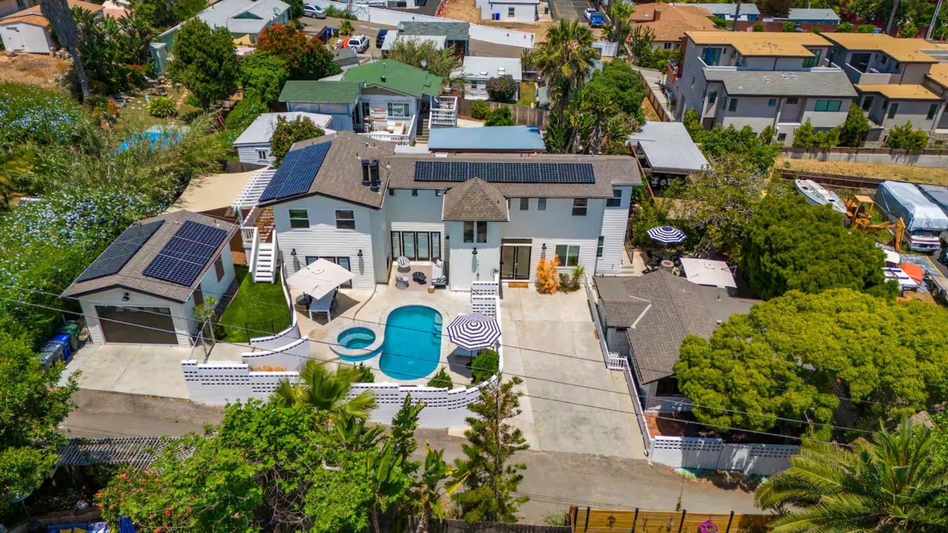 Aerial view of a backyard with a swimming pool and hot tub, patio furniture with umbrellas, surrounded by a white fence, neighboring houses, trees, and solar panels on rooftops.