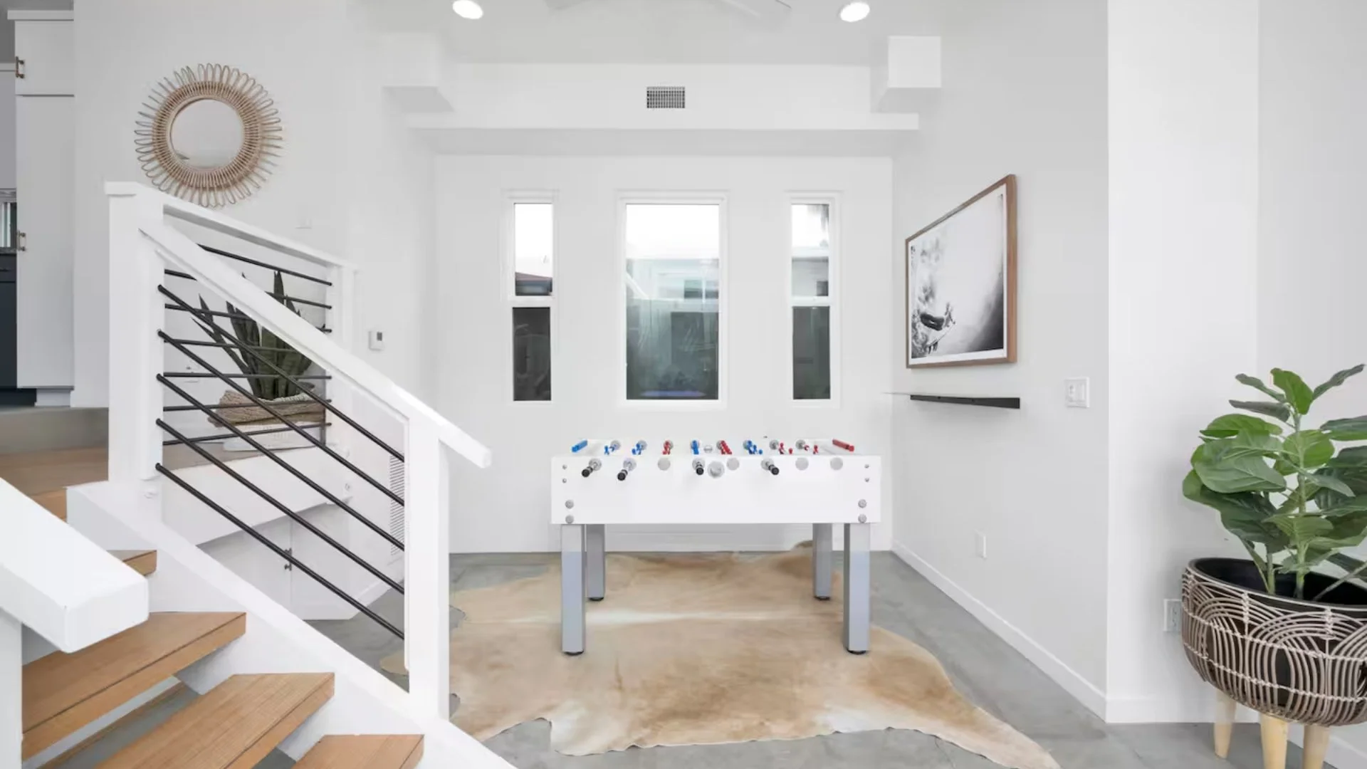 Modern living room with white walls, large windows, a foosball table, indoor plants, wall art, and a cowhide rug.