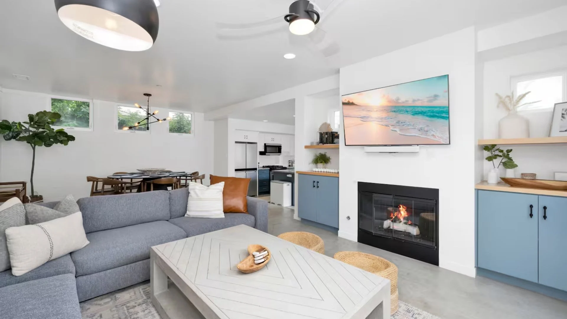 Living room with gray sofa, white coffee table, blue cabinets, fireplace, wall-mounted TV showing a beach scene, and dining area with wooden table and chairs in the background.