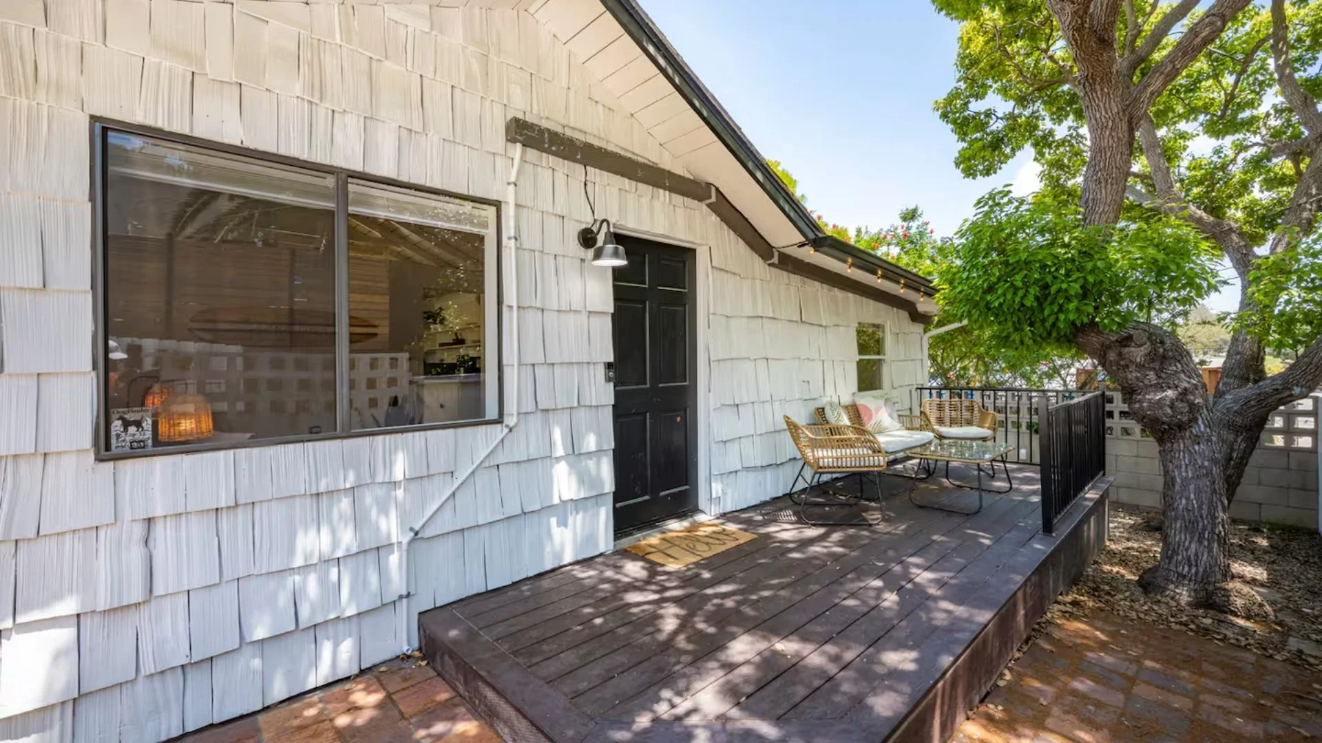 A small outdoor balcony with wicker furniture and cushions, surrounded by a tree with lush green leaves, attached to a house with white shingle siding and a large window.