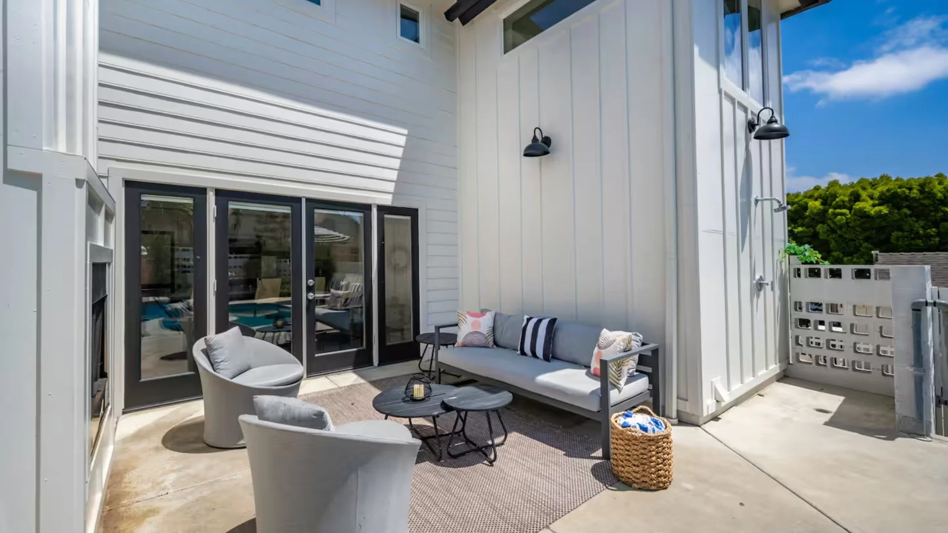 Outdoor patio with modern furniture, including two white chairs, a gray sofa with patterned pillows, and black nesting tables, on a textured rug. There are two outdoor wall lights, a woven basket with blankets, and sliding glass doors leading into a 