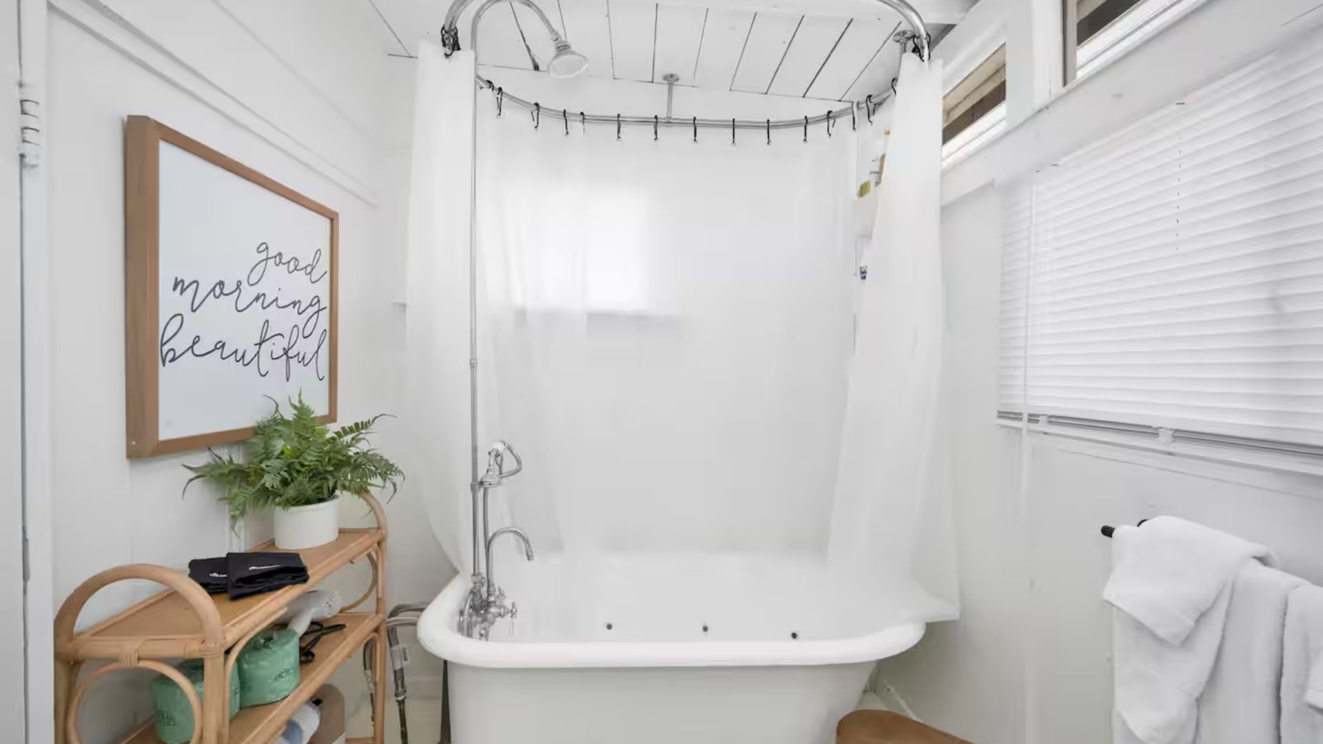 A bright bathroom with a clawfoot bathtub, white shower curtain, framed wall art reading 'good morning beautiful,' a wicker shelf with a plant and towels, and window blinds.