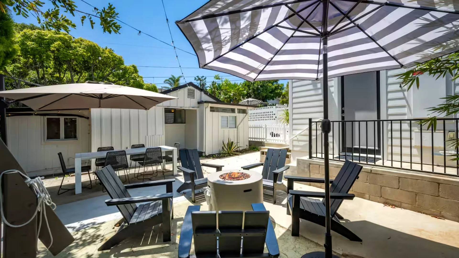 Outdoor patio with black Adirondack chairs arranged in a circle around a fire pit, shaded by a large striped umbrella. There is a white dining table with chairs under another umbrella, a white shed, some greenery, and string lights overhead.